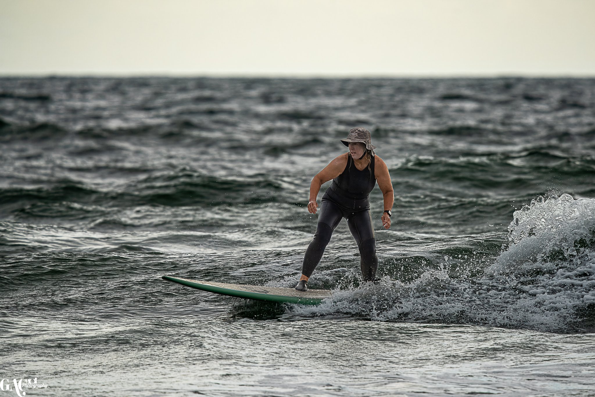 Woman surfing on a surfboard in the ocean, wearing a hat and black wetsuit.
