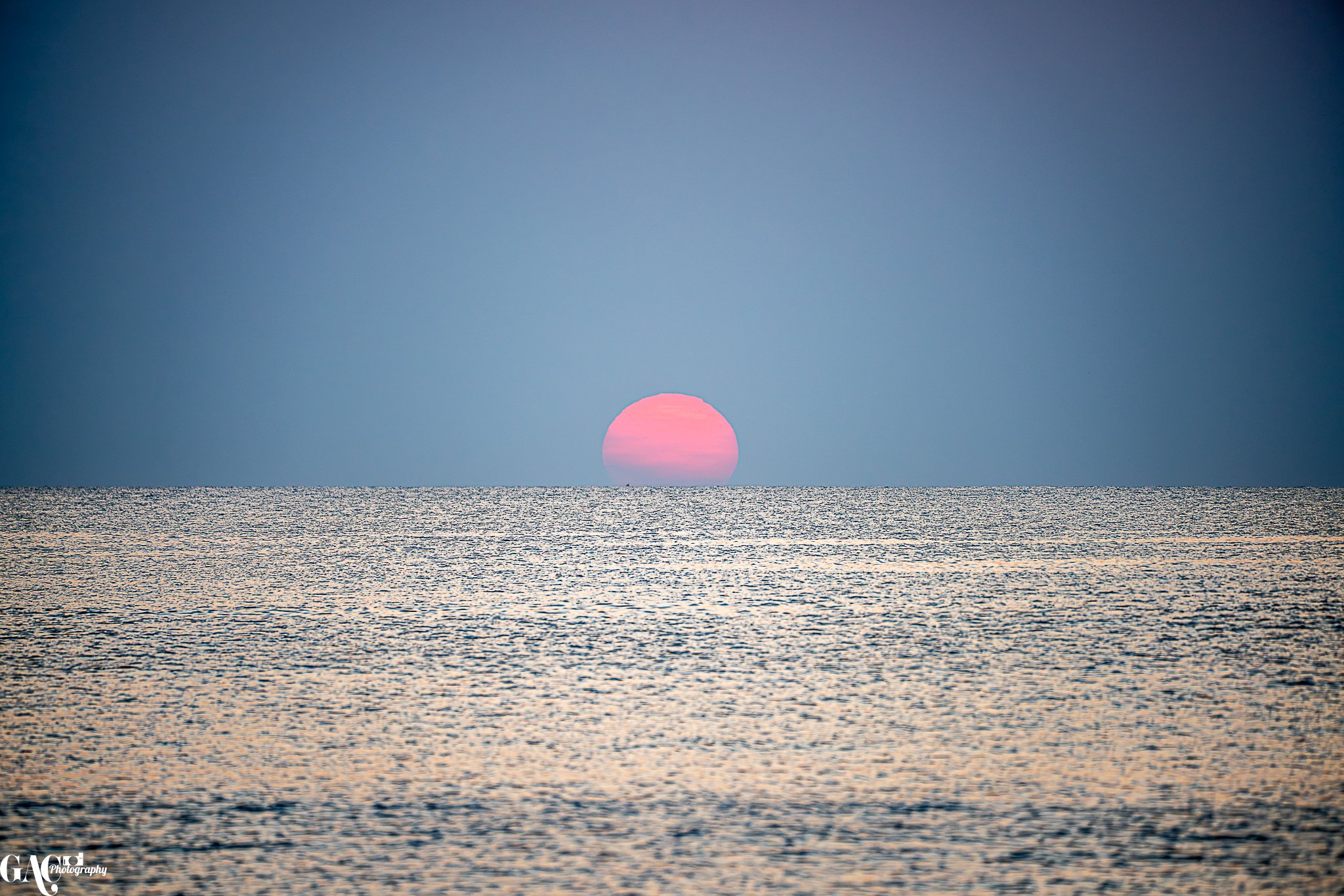 A photograph of a sunset over the ocean showing a pink sun near the horizon with a calm sea and a clear sky.