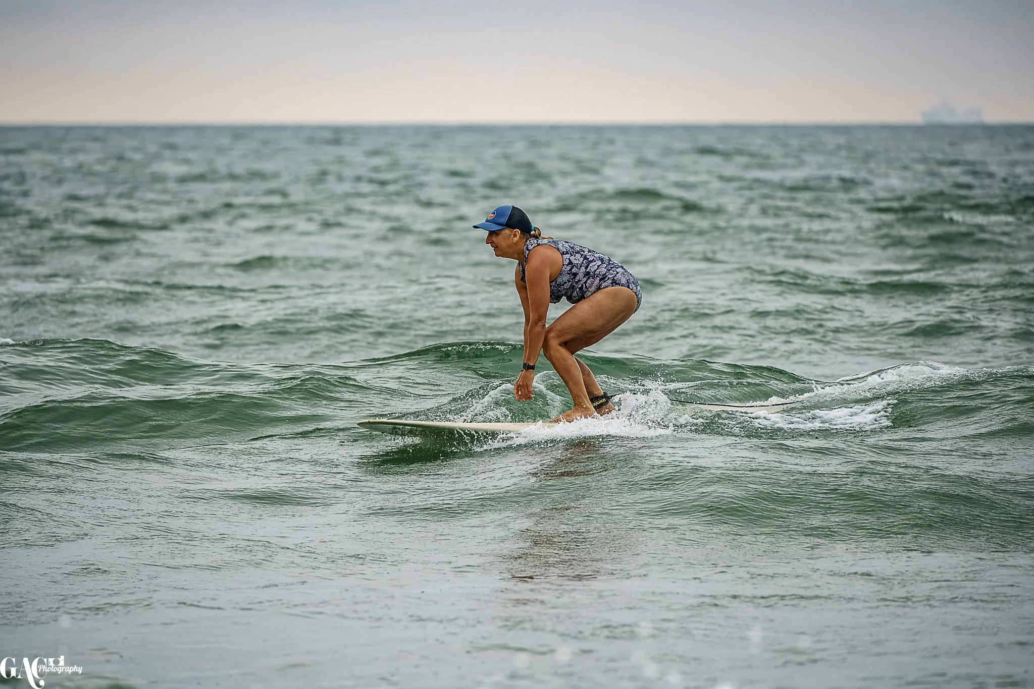 Woman surfing on the ocean, wearing a blue cap, patterned swimsuit, and black ankle strap, crouched on her surfboard.