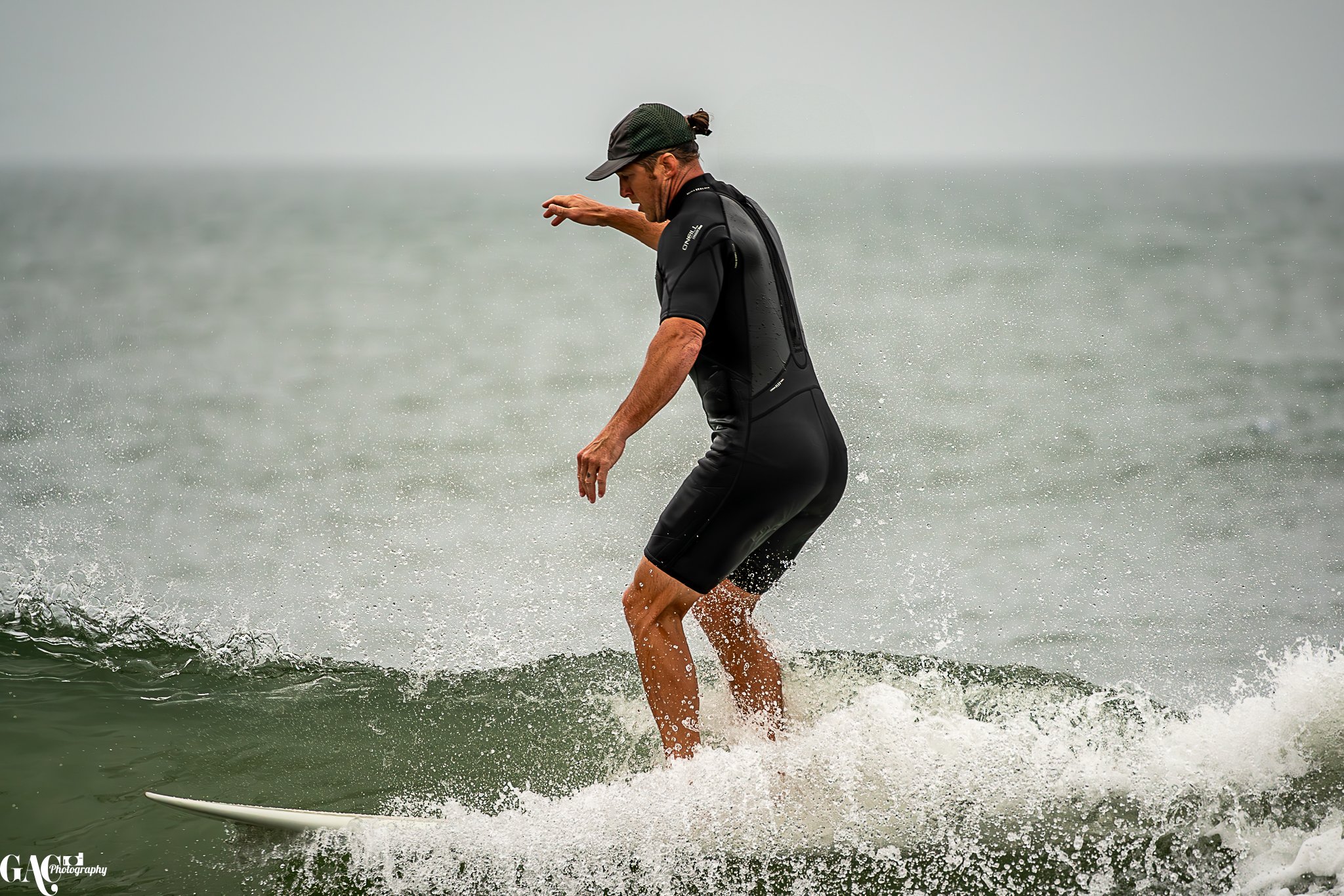 A man wearing a black cap, black wetsuit, and riding a surfboard on a small wave in the ocean on a cloudy day.