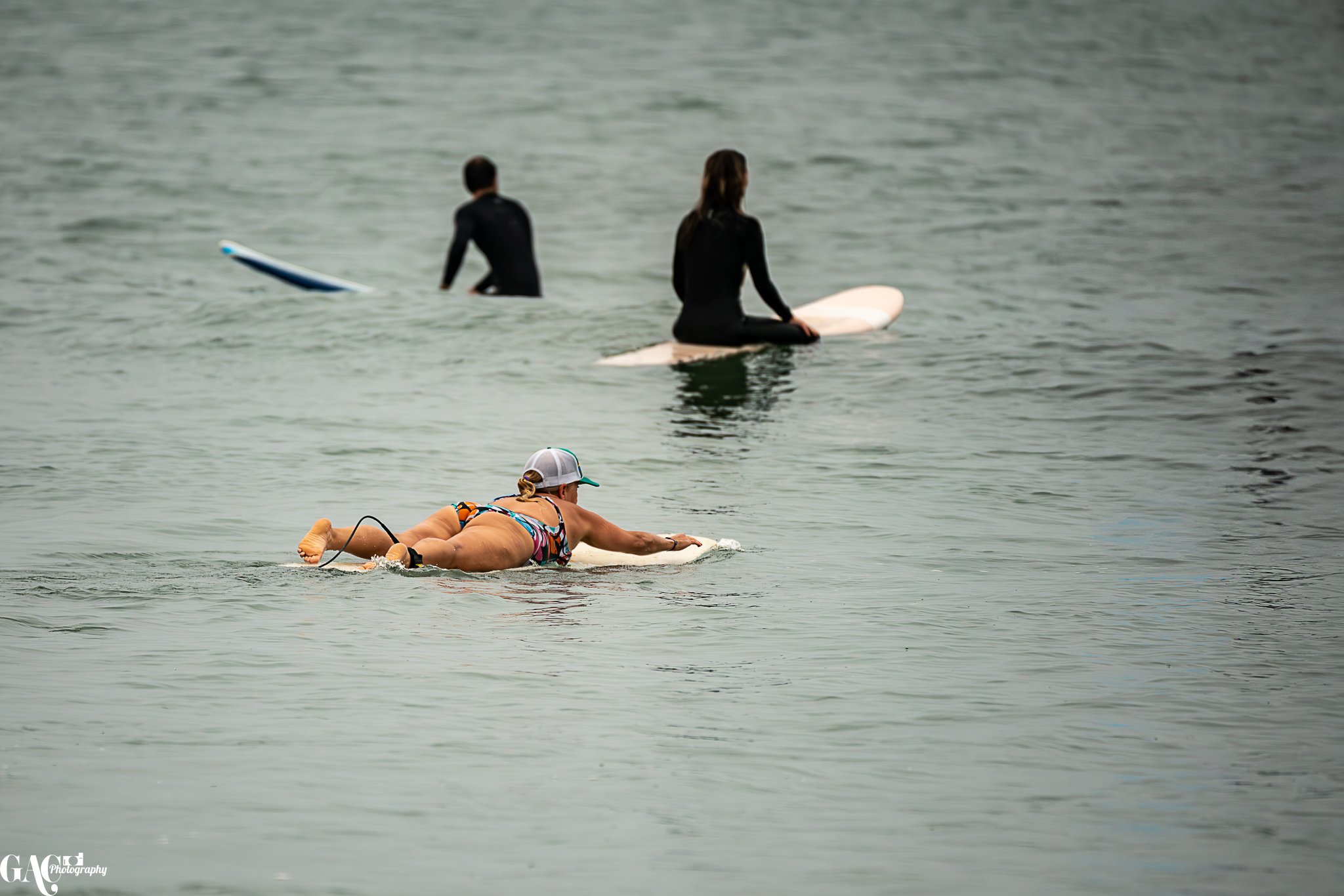 A woman in a swimsuit and cap lying on a surfboard in the ocean, with two people in wetsuits sitting on surfboards in the background.