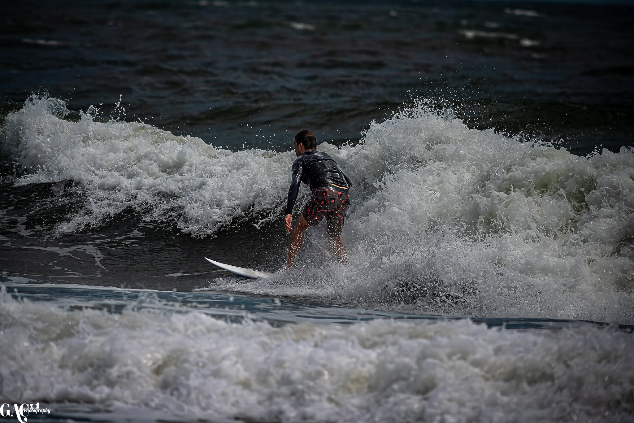 A person surfing on a wave in the ocean, wearing a black wetsuit and swim shorts with a colorful pattern.