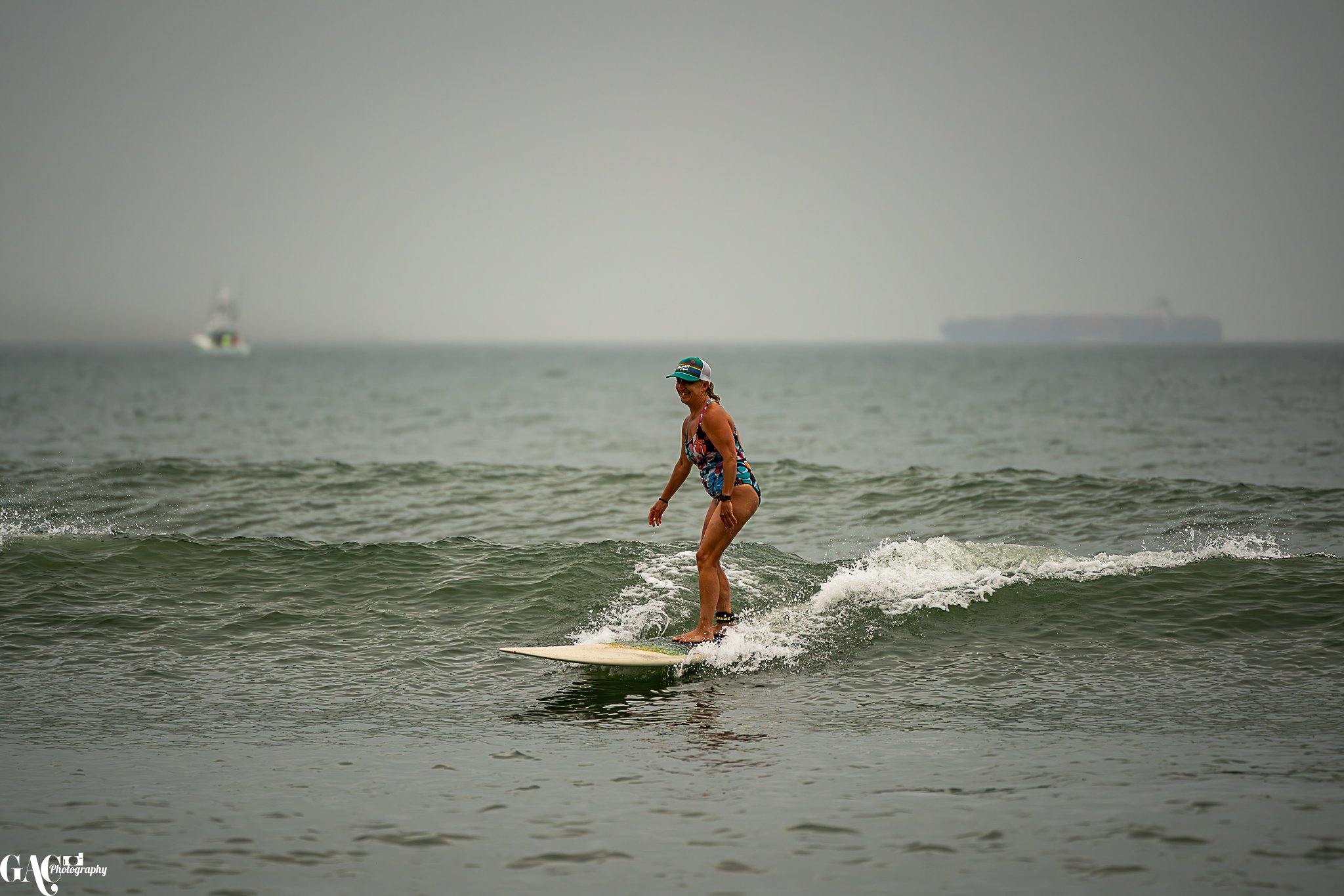 Woman surfing on a small wave in the ocean, wearing a colorful swimsuit and a cap, with ships in the background.