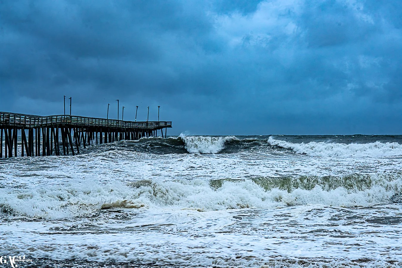 Stormy ocean waves crashing against a wooden pier under a dark cloudy sky.