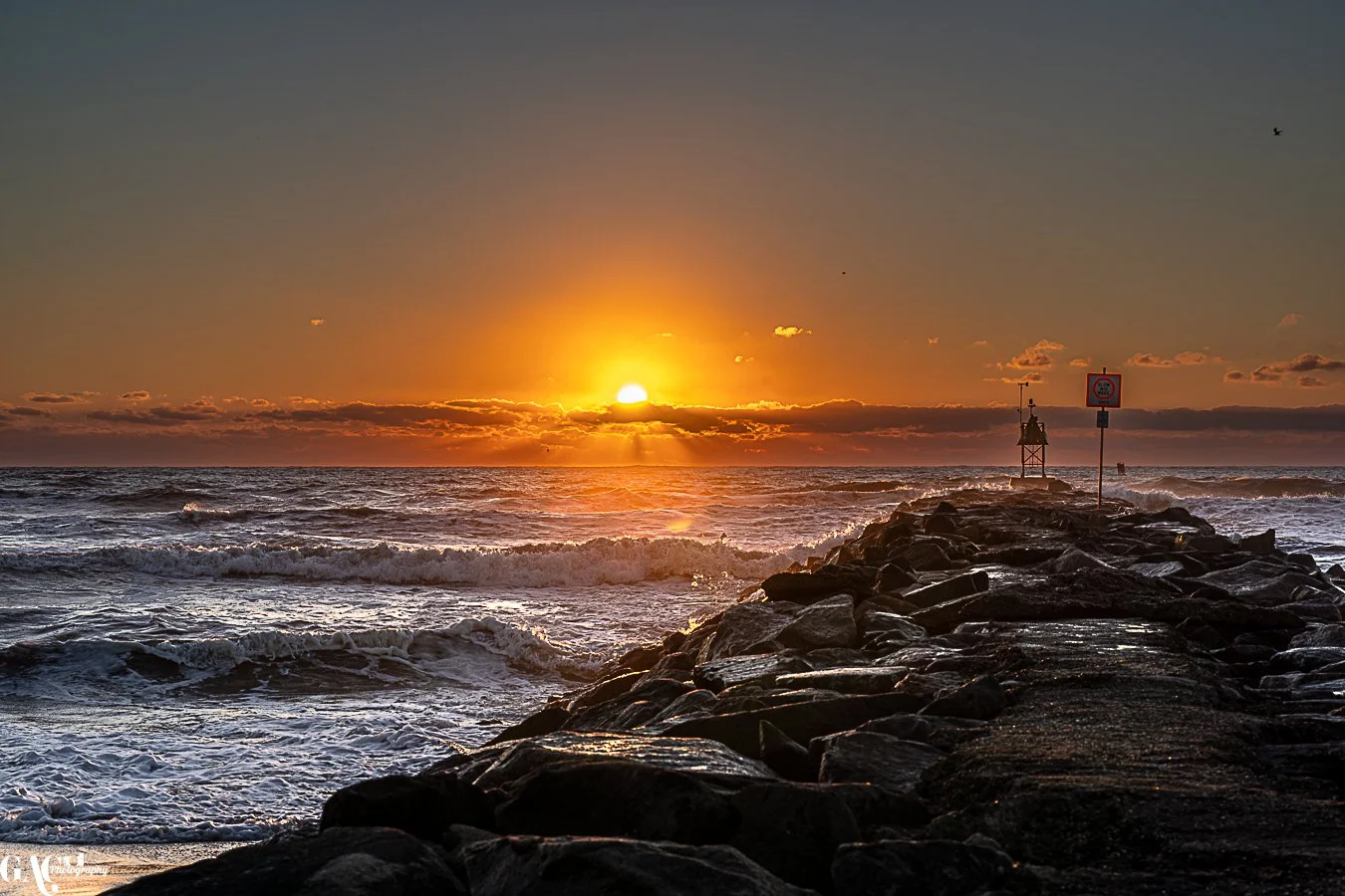 Sunset over the ocean with waves crashing near a rocky pier, lighthouse at the end, and a signpost