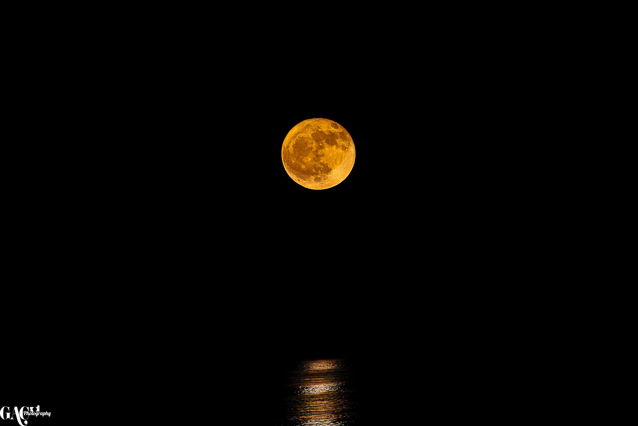 Full moon with yellow-orange hue rising above a dark horizon, reflecting on water surface at night.