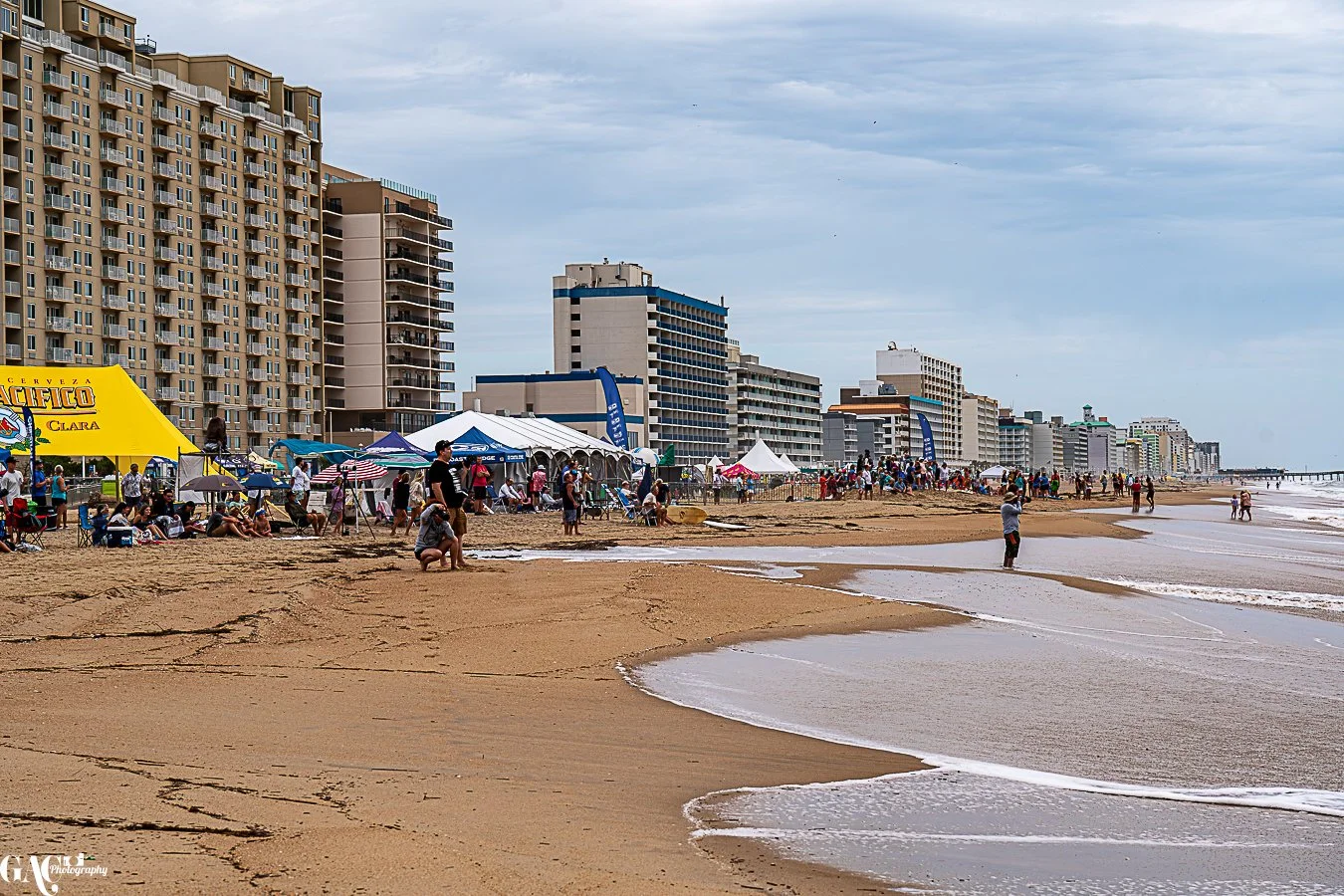 Beach with high-rise buildings along the shoreline, people sitting under umbrellas, tents, and walking near the water on a cloudy day.