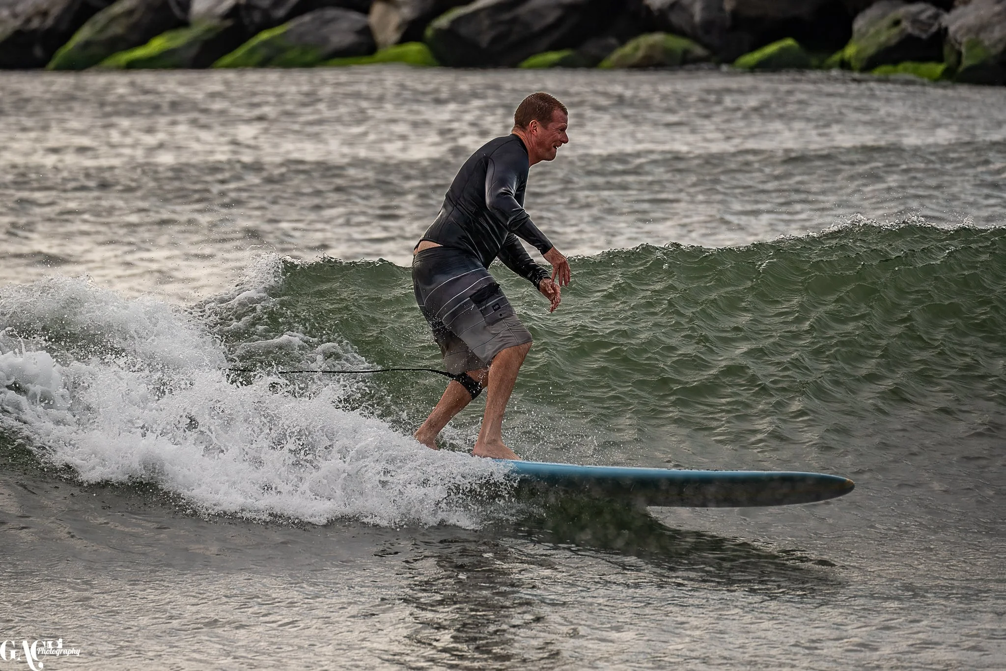 Man surfing on a wave, wearing a black wetsuit and shorts, in the water.