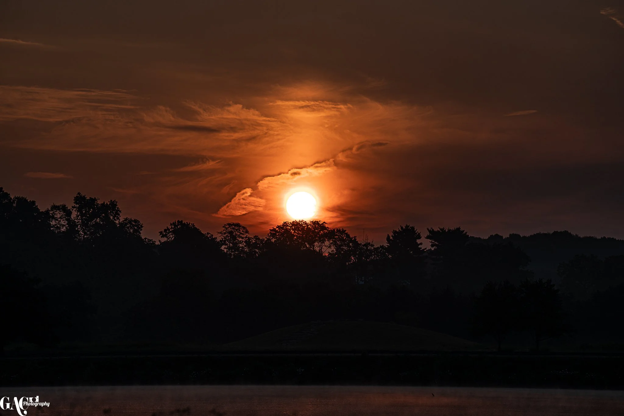 Sunset over a wooded landscape with dark trees silhouetted against an orange sky, with clouds illuminated by the setting sun.