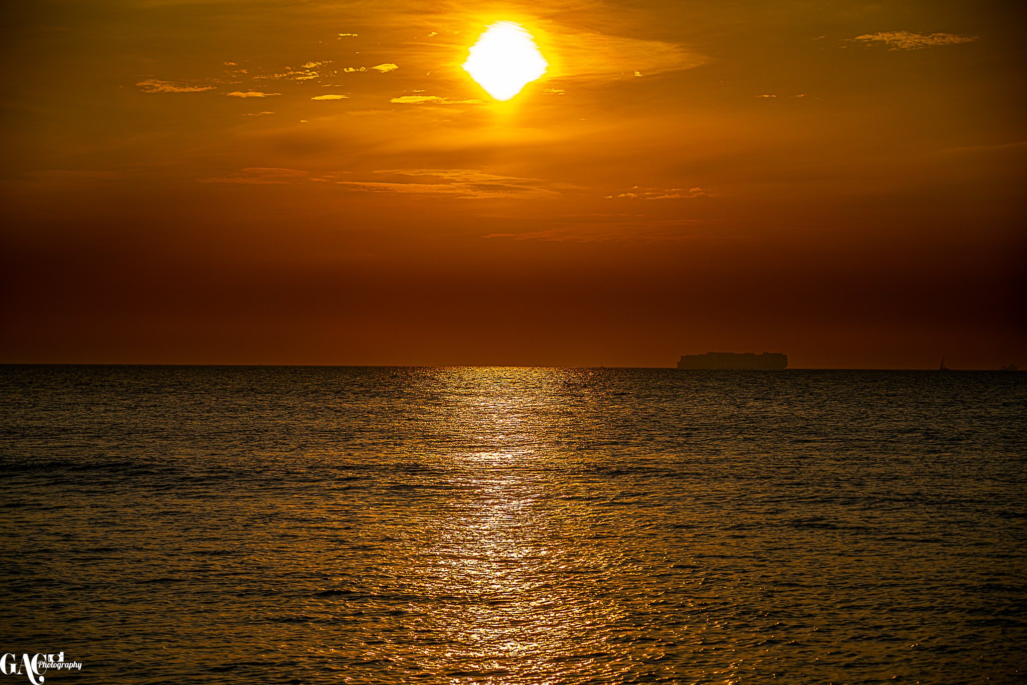 Sunset over the ocean with a shiny reflection on the water, a large cargo ship in the distance, and a partly cloudy sky.
