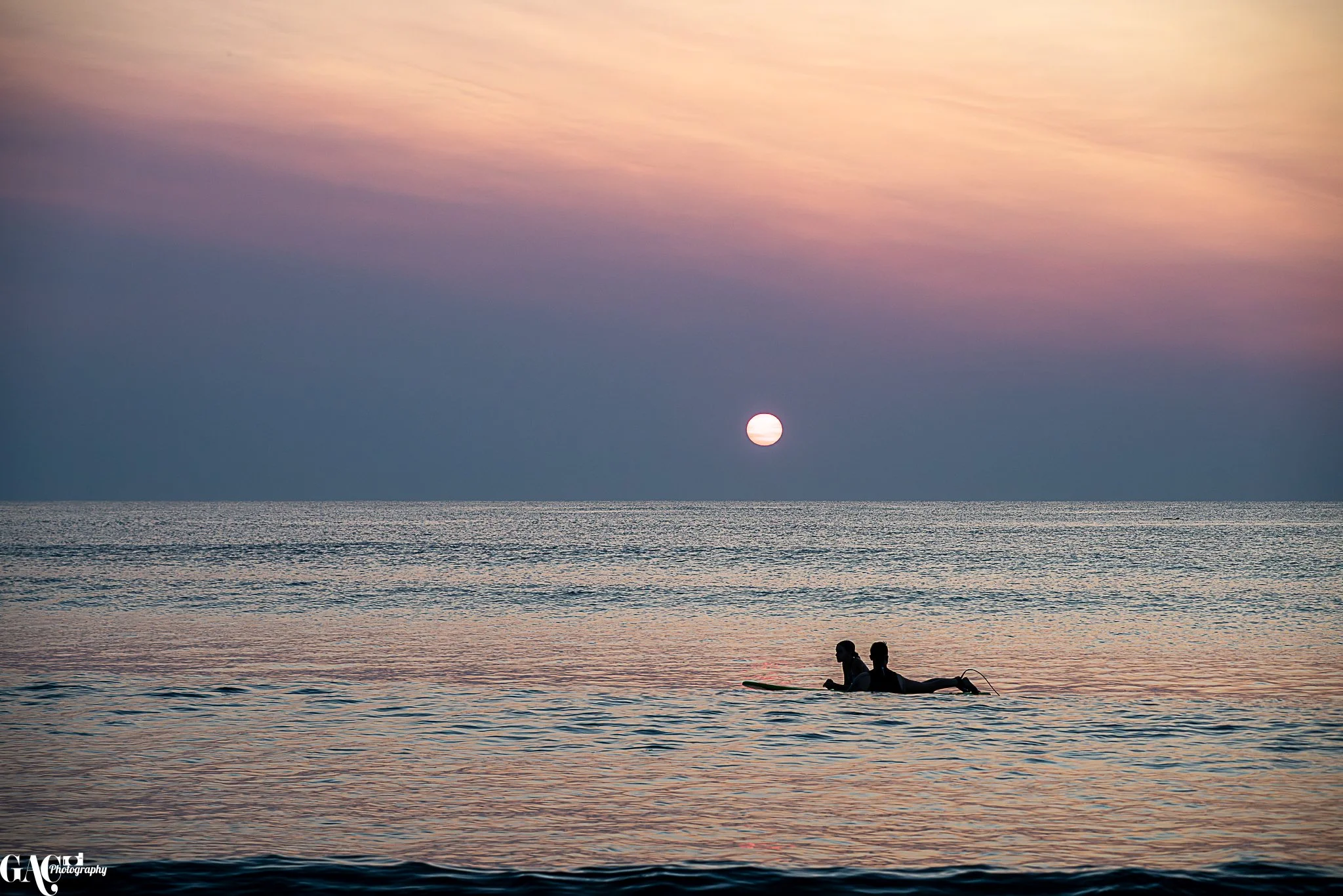 Silhouettes of two people on a surfboard in the ocean during sunset, with colorful sky and the sun near the horizon.