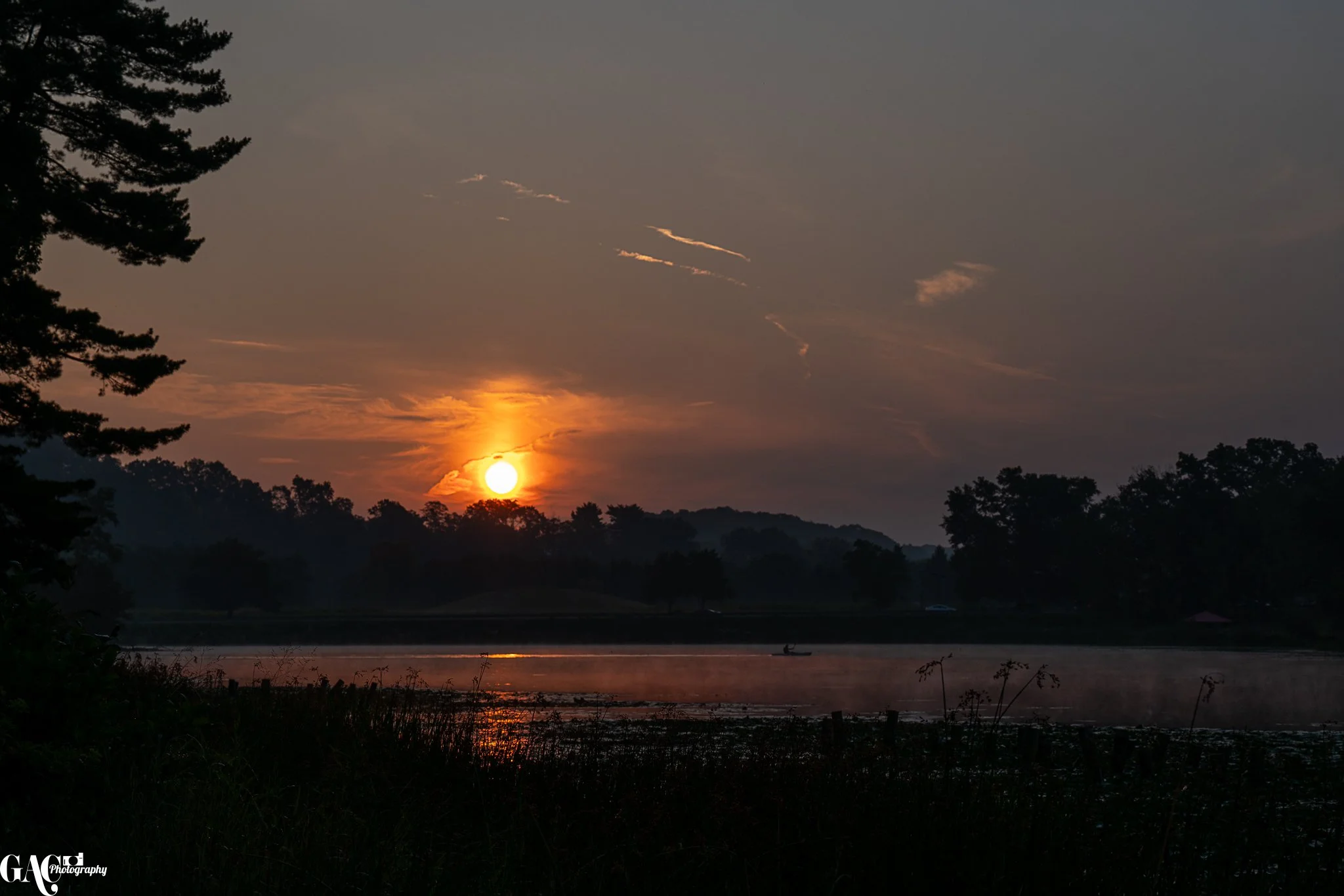 Sunset over a lake with mist rising from the water, surrounded by trees and a hilly landscape in the distance.