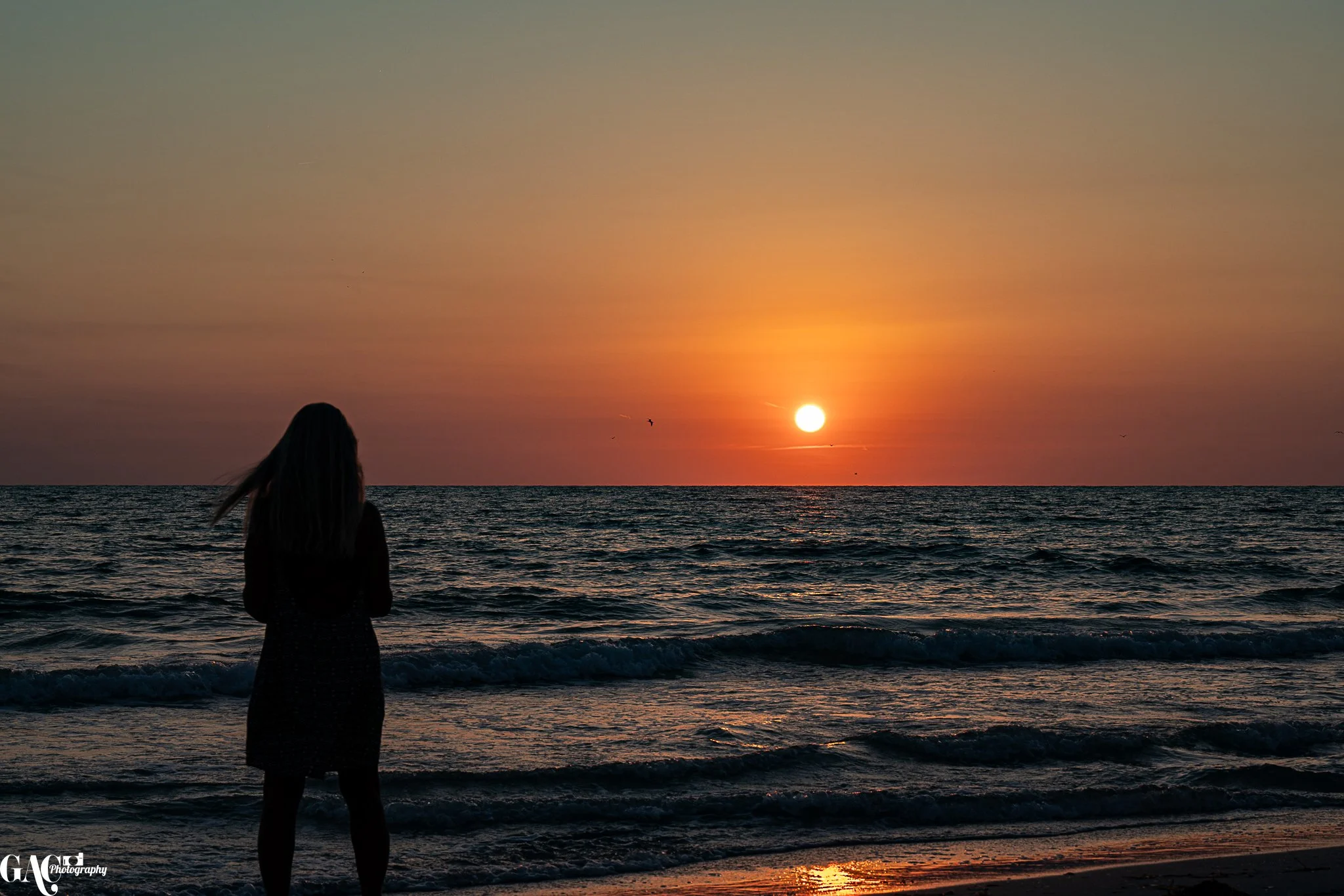 A woman standing on the beach during sunset, facing the ocean with the sun low on the horizon, in silhouette.