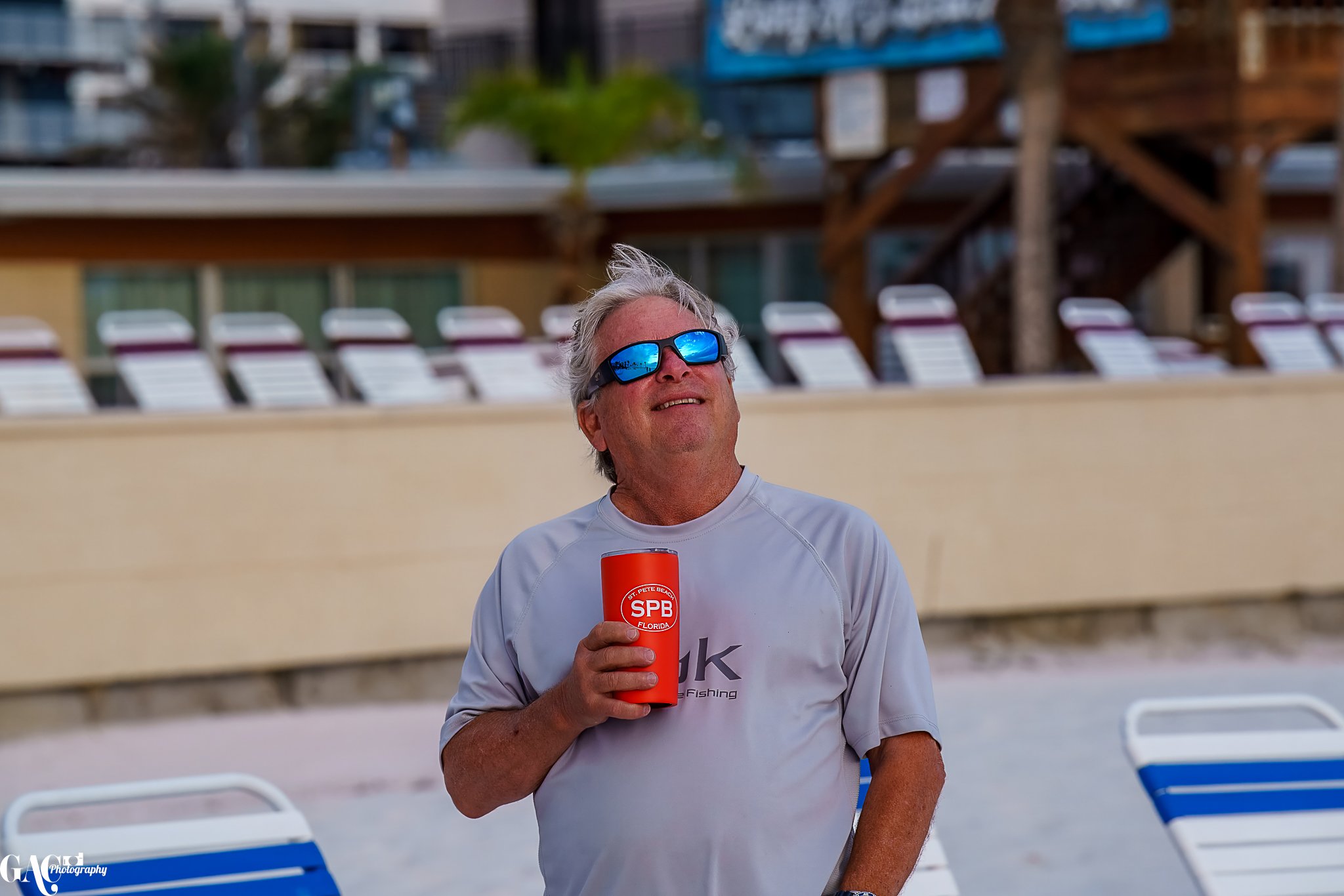 Man wearing sunglasses holding SPB Florida drink tumbler at the beach, with lounge chairs in the background.