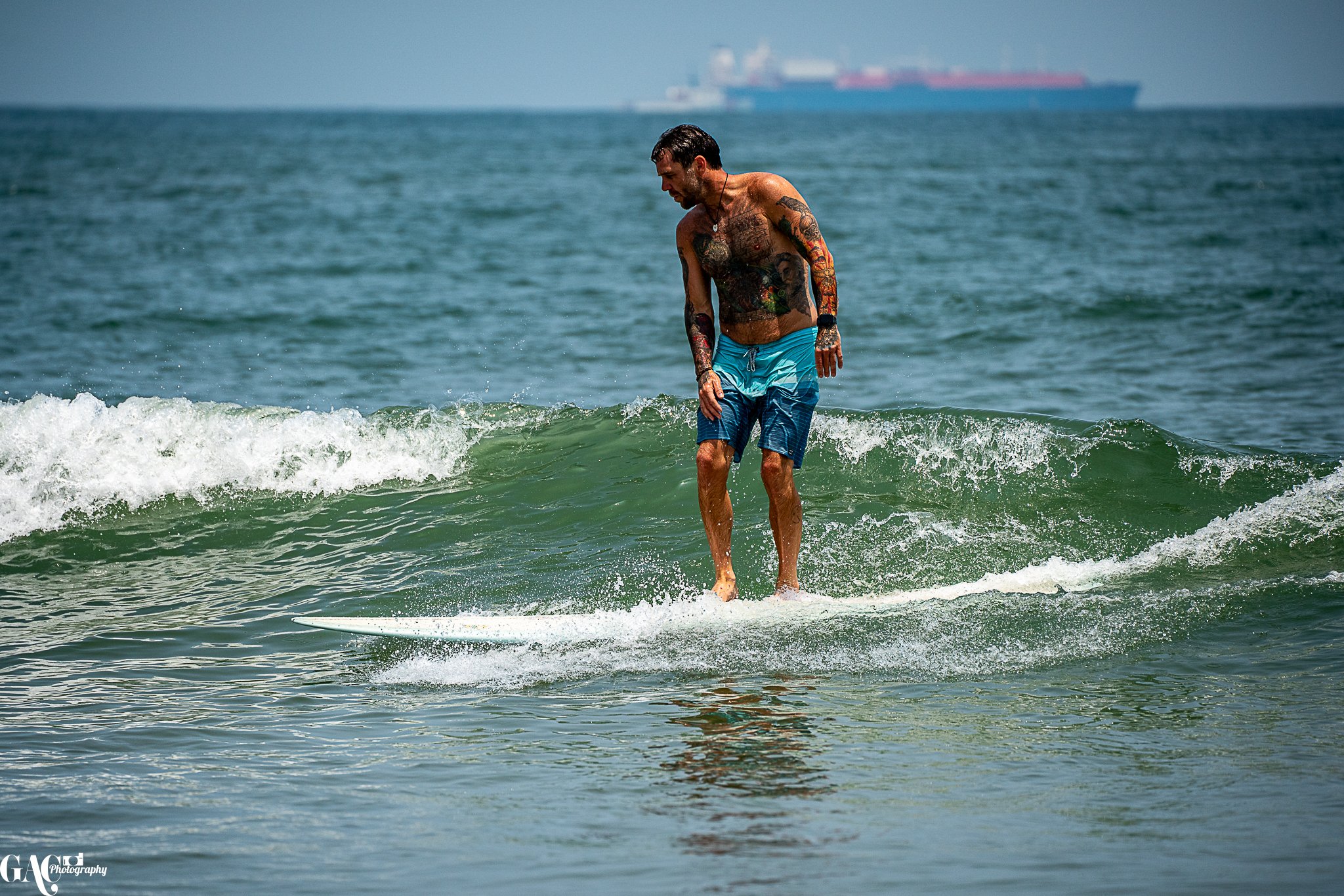 A tattooed man in blue swim shorts surfing on a small wave at the beach with a large cargo ship visible in the distance.