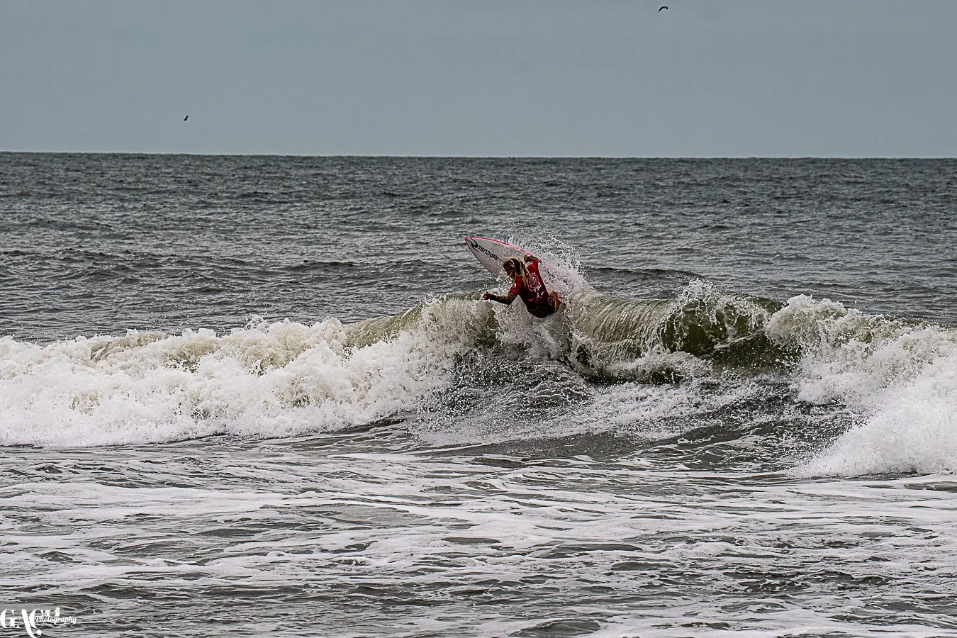 A person in a red wetsuit riding a surfboard on a breaking wave at the beach.