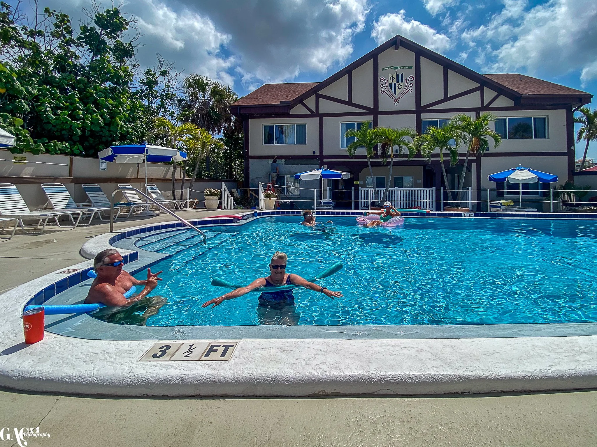 Outdoor swimming pool with people relaxing and using pool noodles, surrounded by lounge chairs and umbrellas, with a building in the background.