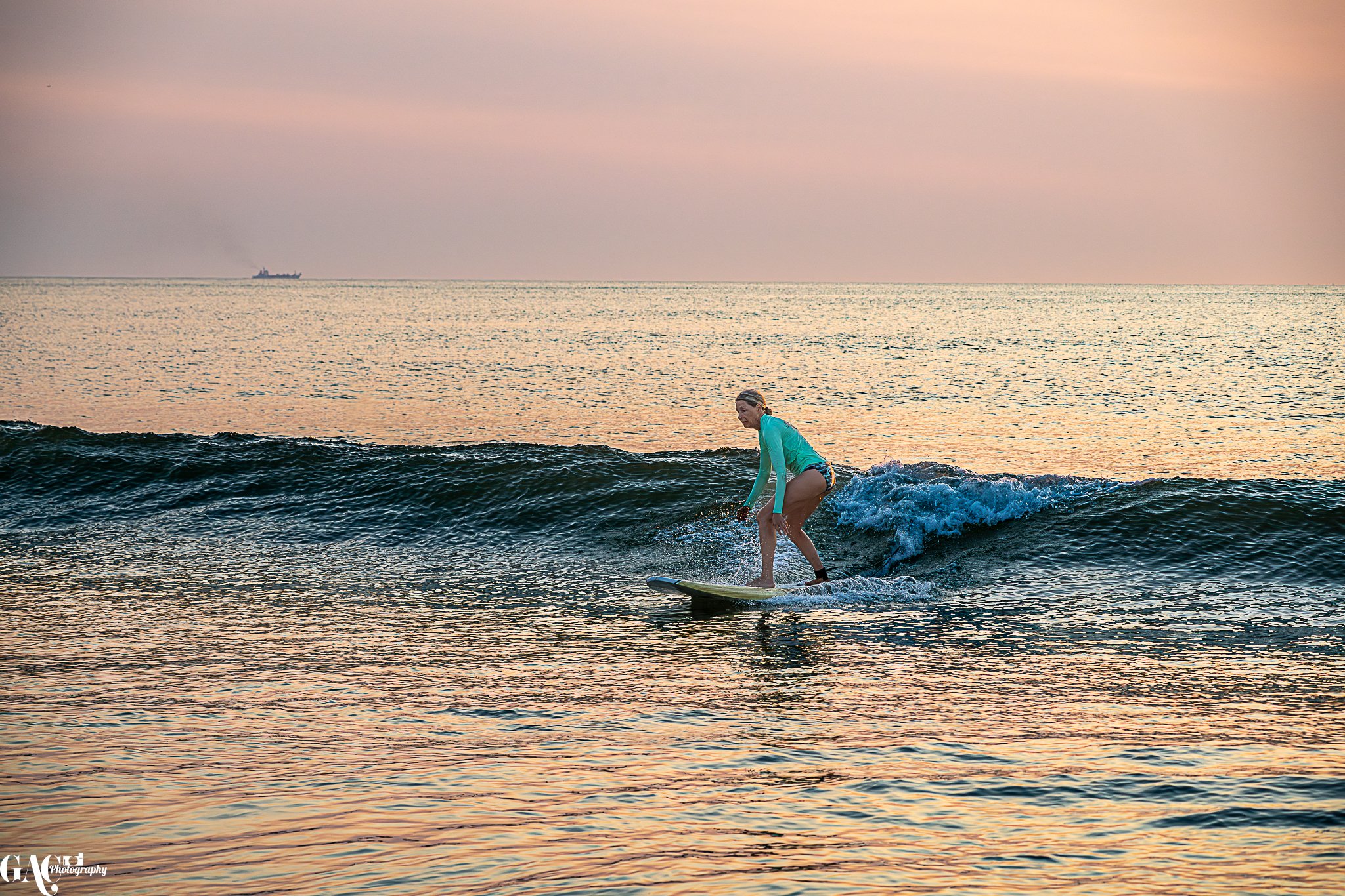 A woman surfing on a small wave during sunset, wearing a turquoise long-sleeve top and shorts.