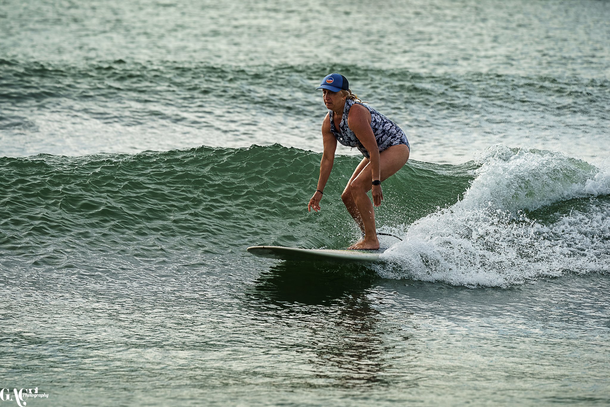 A woman surfing on a small wave in the ocean, wearing a patterned swimsuit and a blue cap.