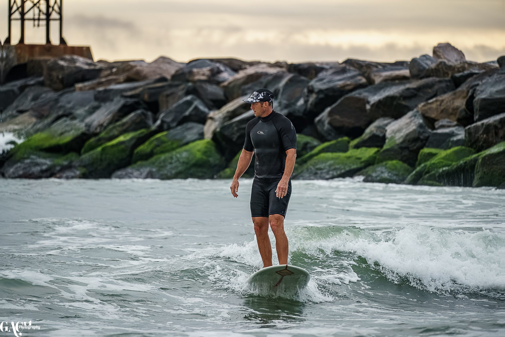 A man surfing in the ocean near rocks covered in green algae, with a cloudy sky in the background.