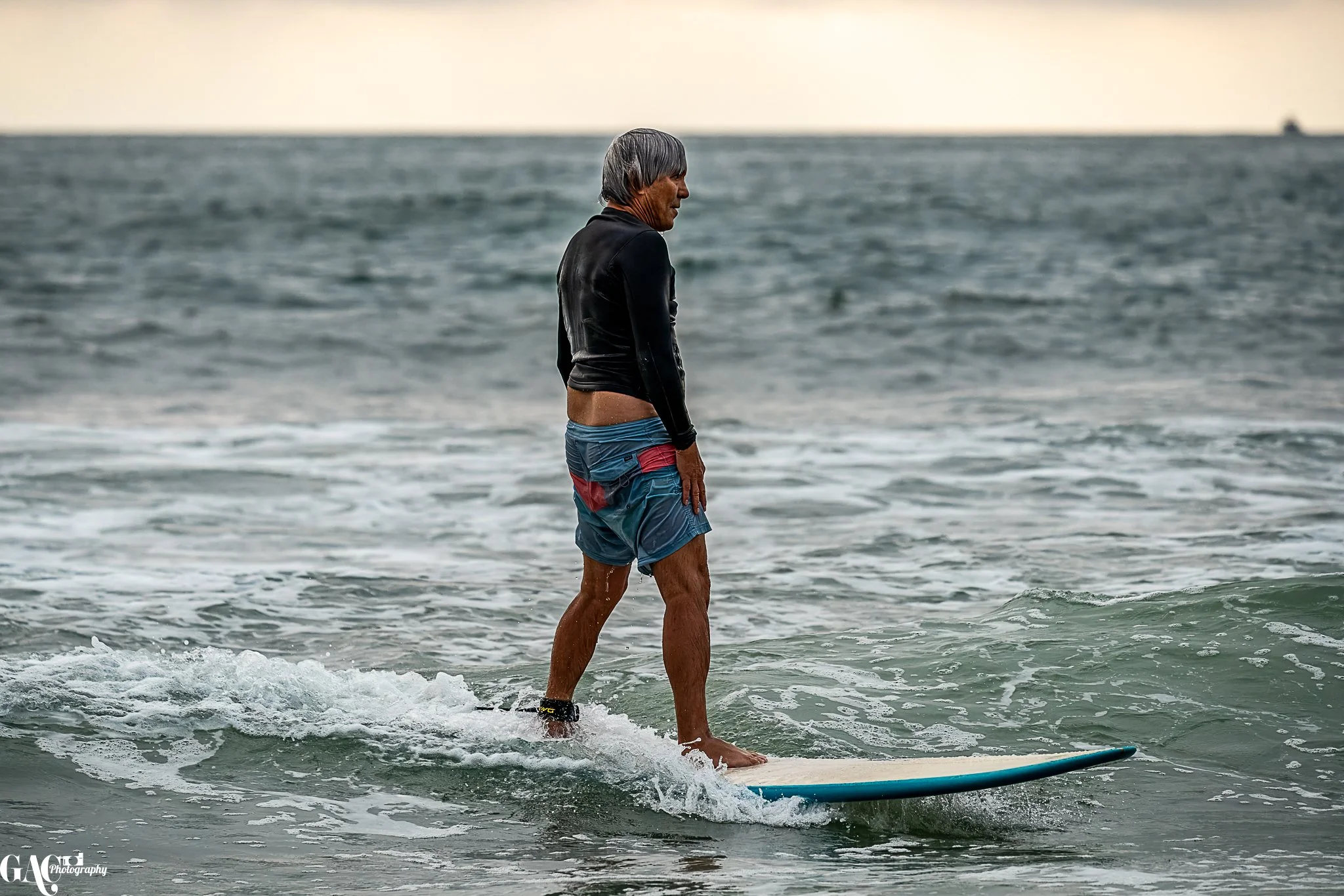 A man standing on a surfboard in the ocean, facing sideways, wearing a black wetsuit top and colorful swim shorts, with gray hair.