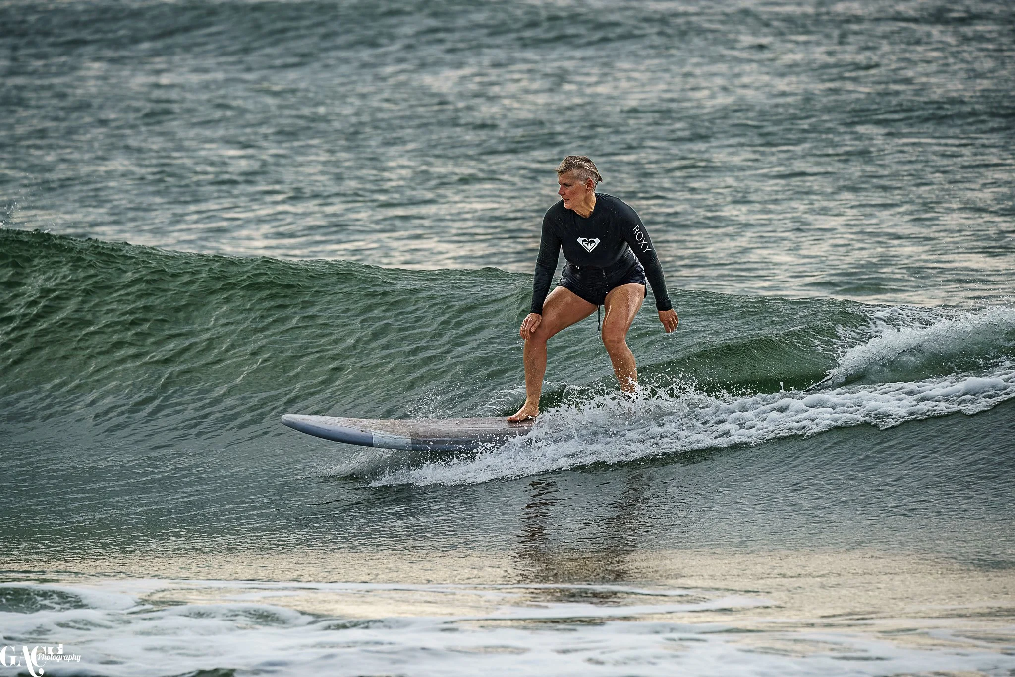 An elderly woman surfing on a small wave in the ocean, wearing a black long-sleeve surf shirt and shorts.