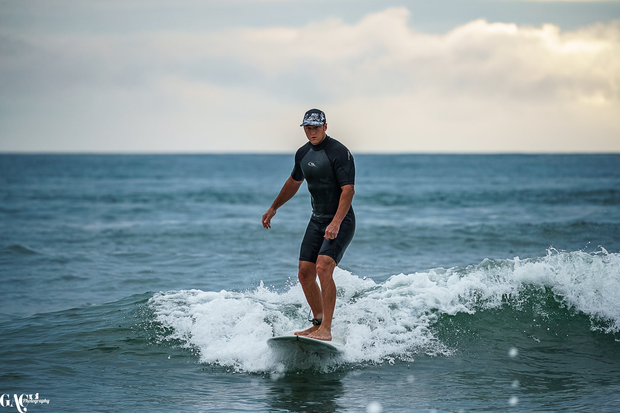 A man surfing on a small wave in the ocean, wearing a black wetsuit and a cap, with a serious expression.