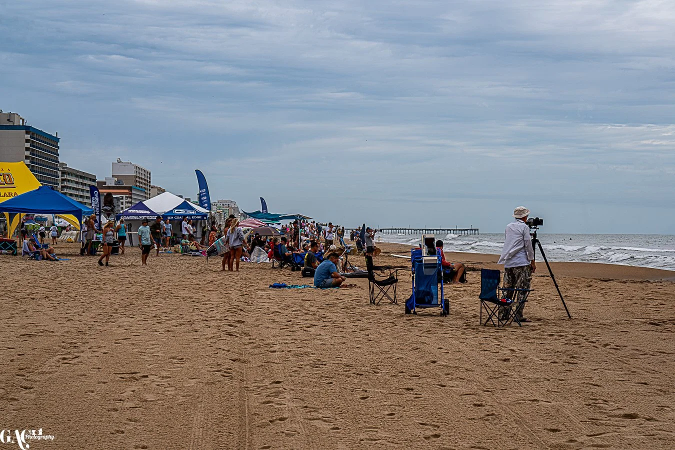 People enjoying a cloudy day at a busy beach with tents, chairs, and a pier in the background.