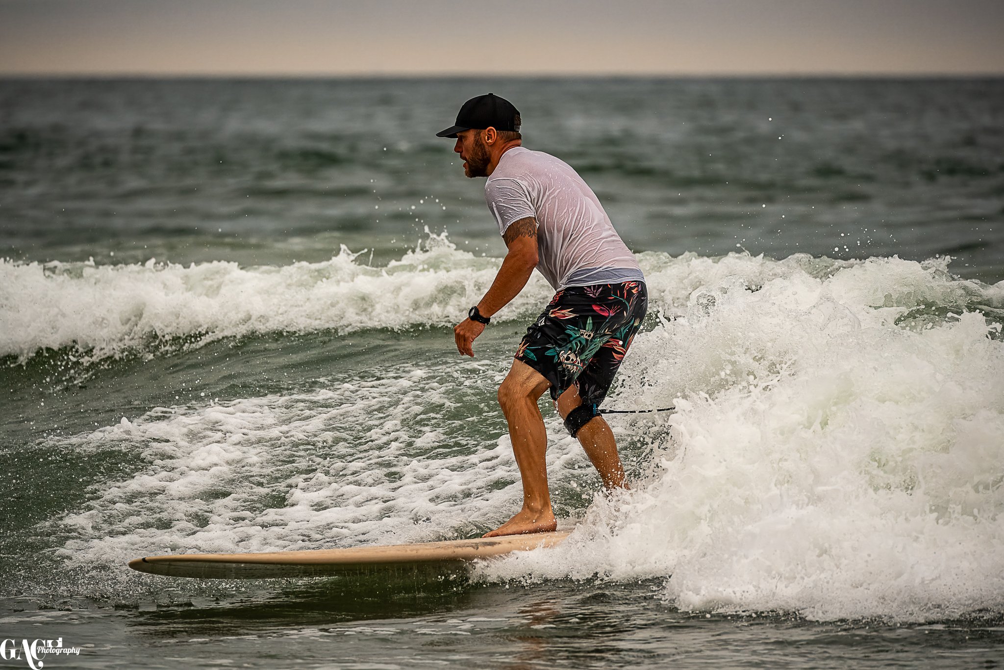 Man surfing on a wave at the beach wearing a gray t-shirt, colorful shorts, and a black cap.