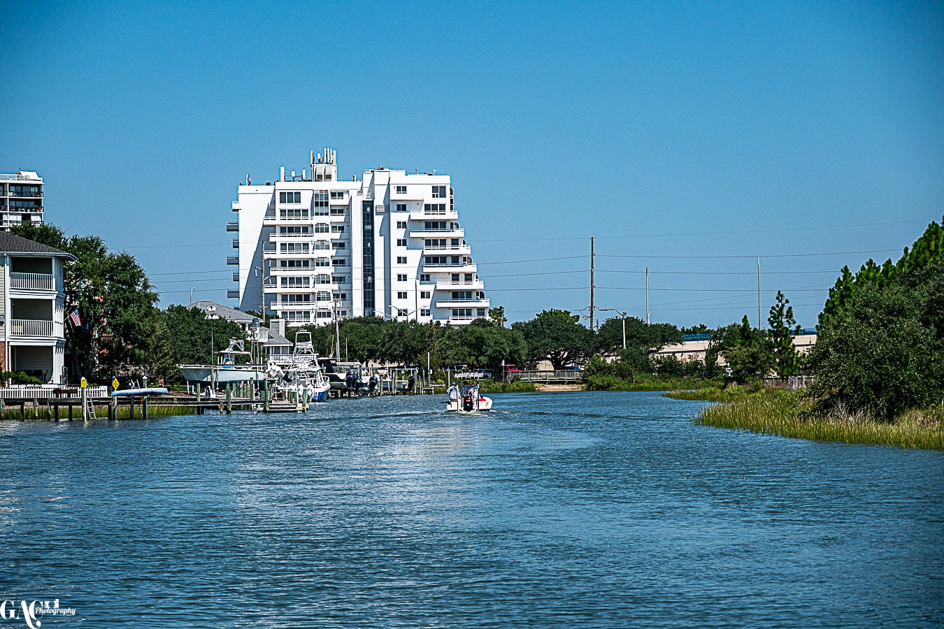 A waterway with boats docked along the sides, greenery on both banks, and a modern white building in the background under a clear blue sky.