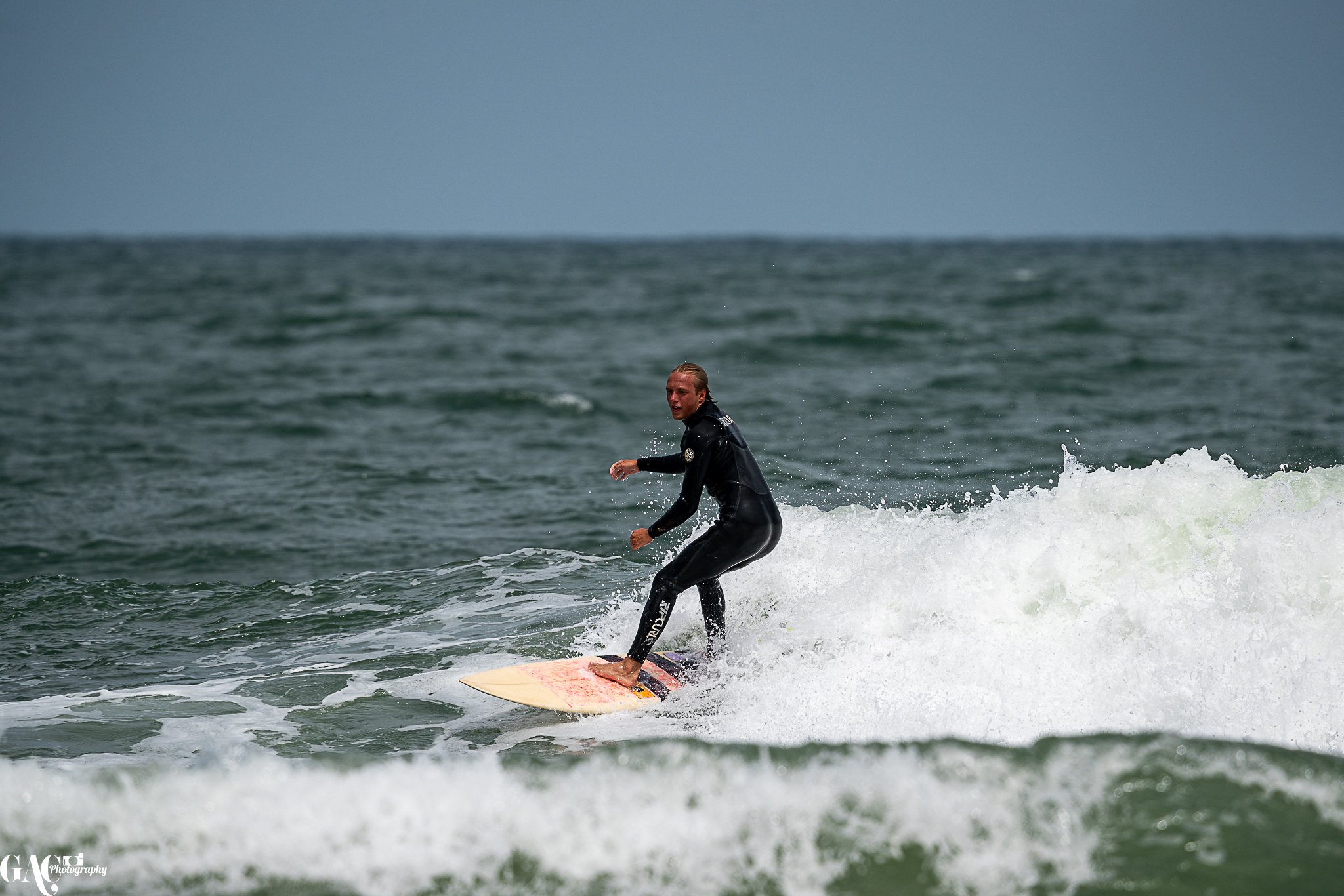 A man surfing in the ocean on a cloudy day, wearing a black wetsuit and riding a white surfboard.