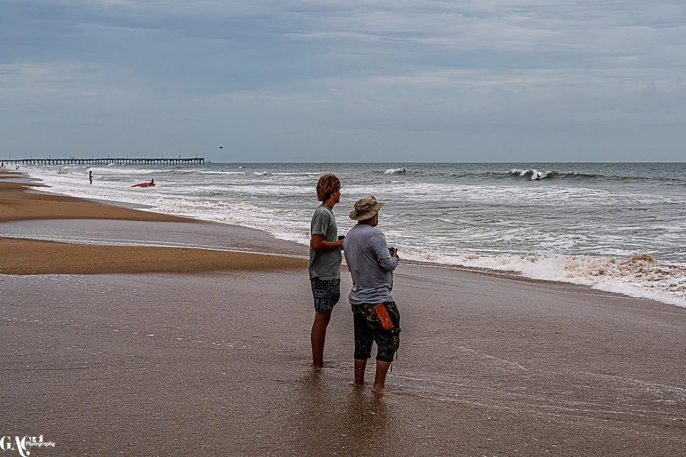 Two people standing at the shoreline on a cloudy day, looking out at the ocean with waves and a pier in the distance.
