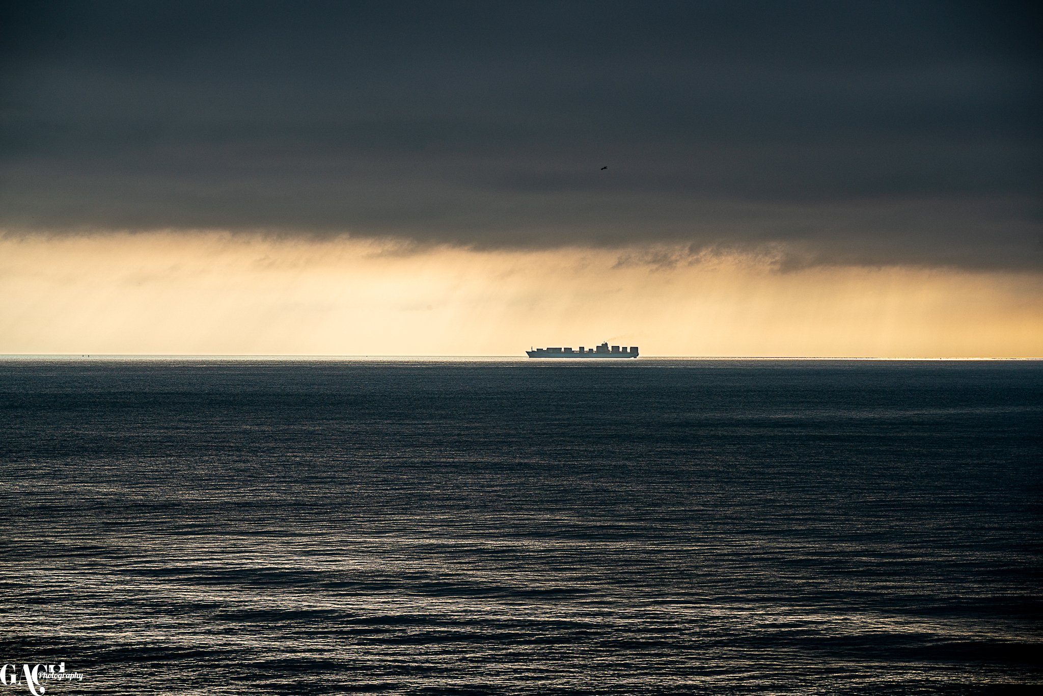 A large cargo ship sailing on the ocean during a cloudy day with dark clouds above and a break in the clouds letting in light, showing a distant horizon.