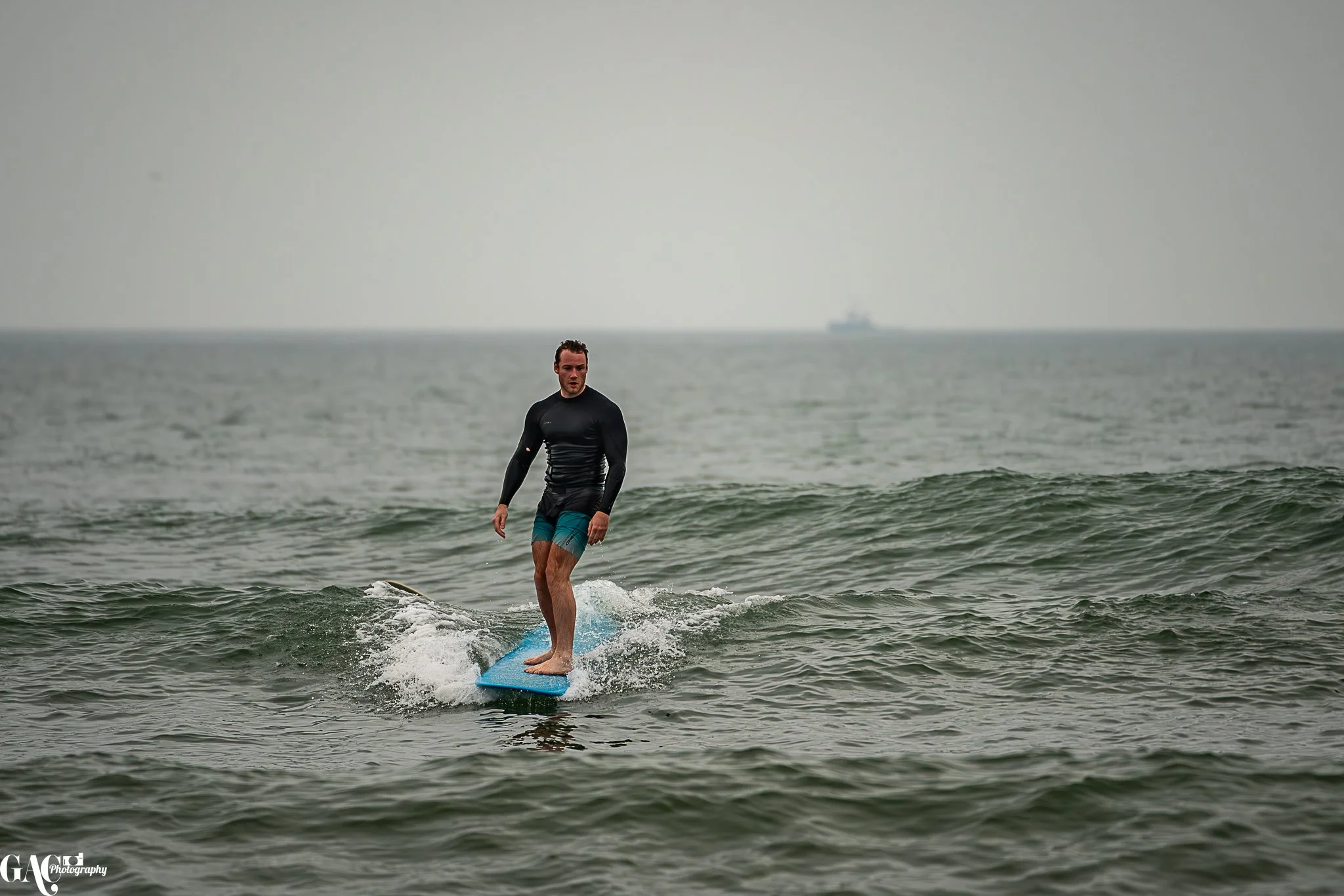 A man in a black long-sleeve shirt and teal shorts is surfing on a blue surfboard in the ocean on a cloudy day.