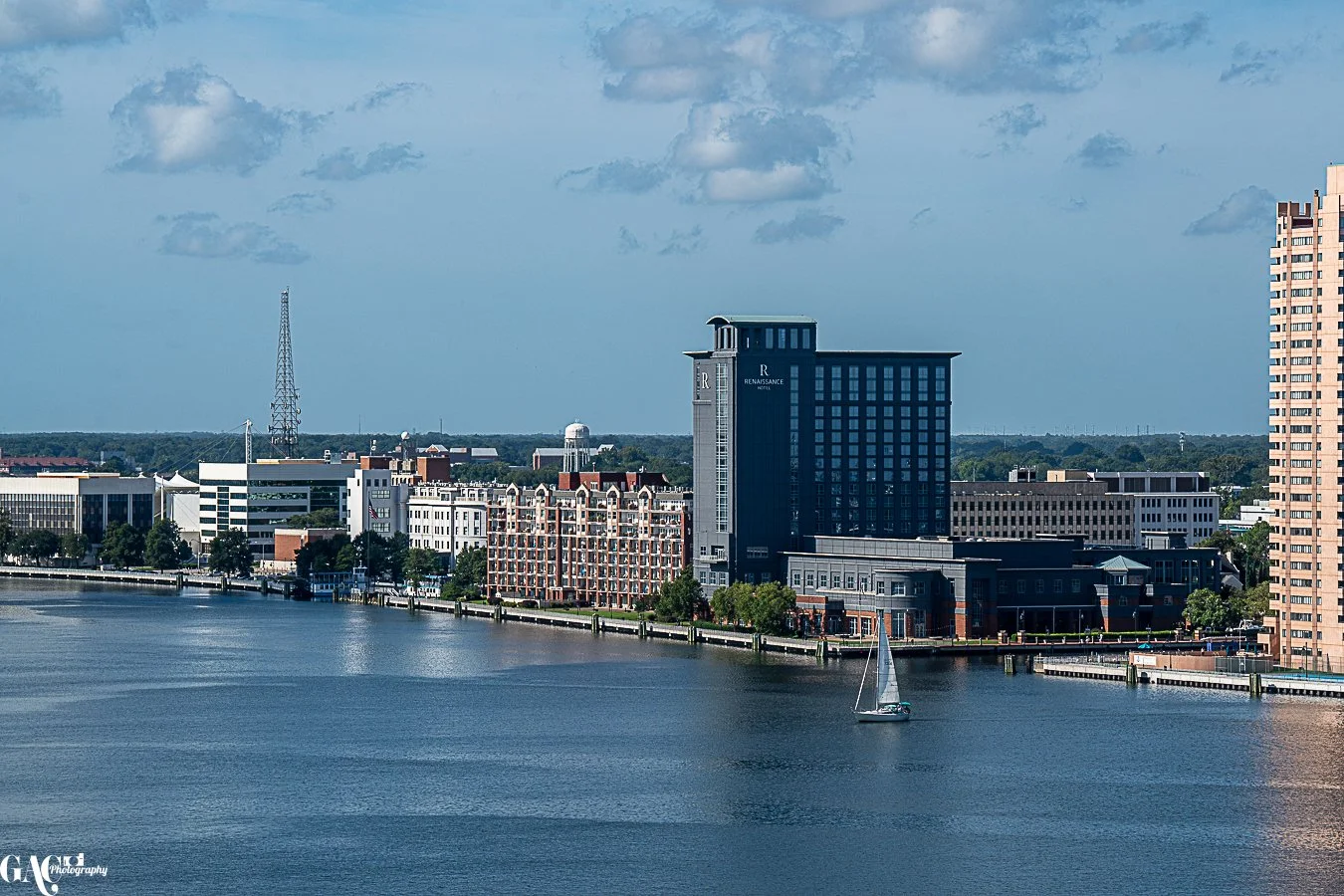 City skyline along a river with a sailboat, high-rise buildings, and a communications tower under a partly cloudy sky.