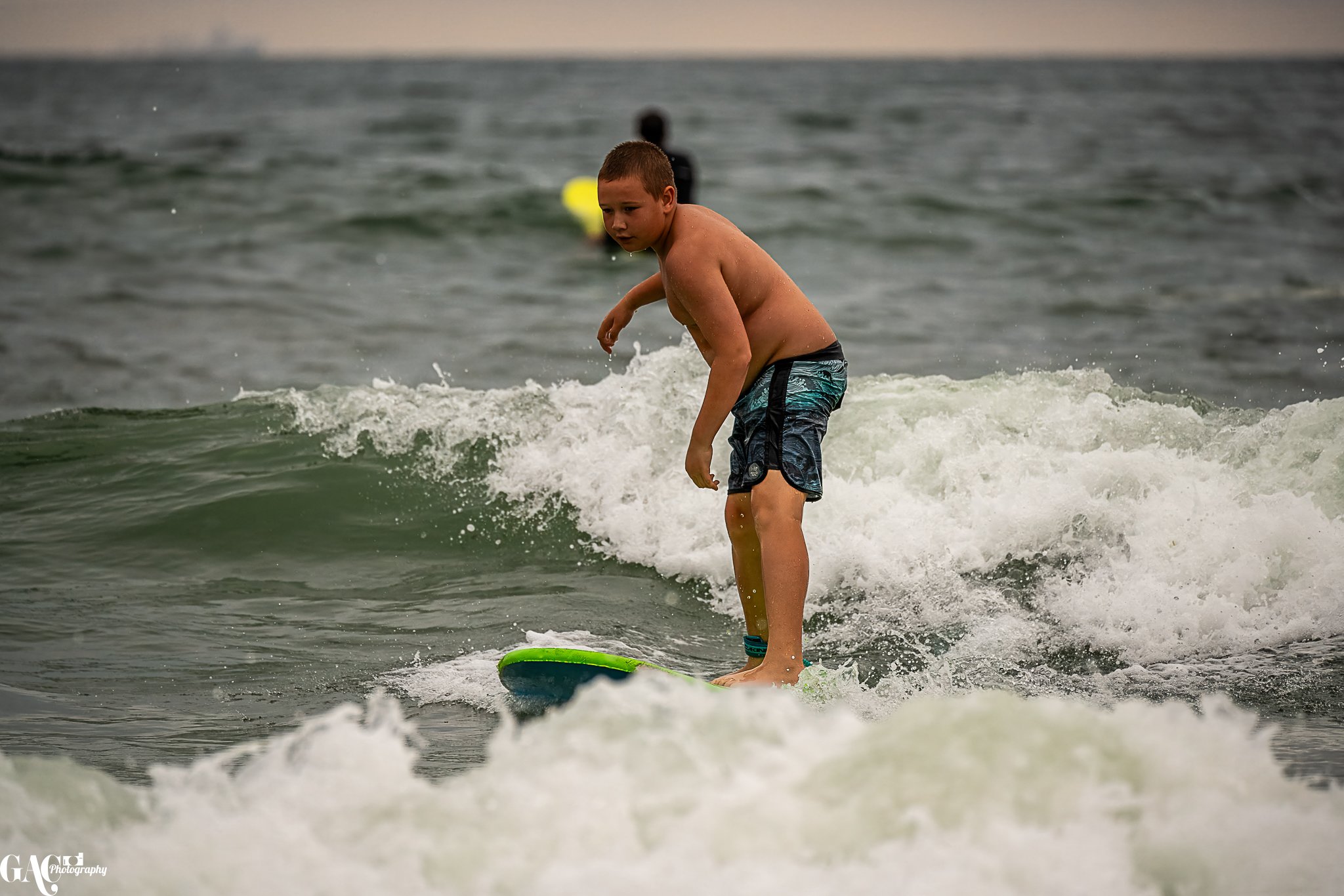 A young boy surfing on a small wave in the ocean, wearing swim shorts, with another person in the background.
