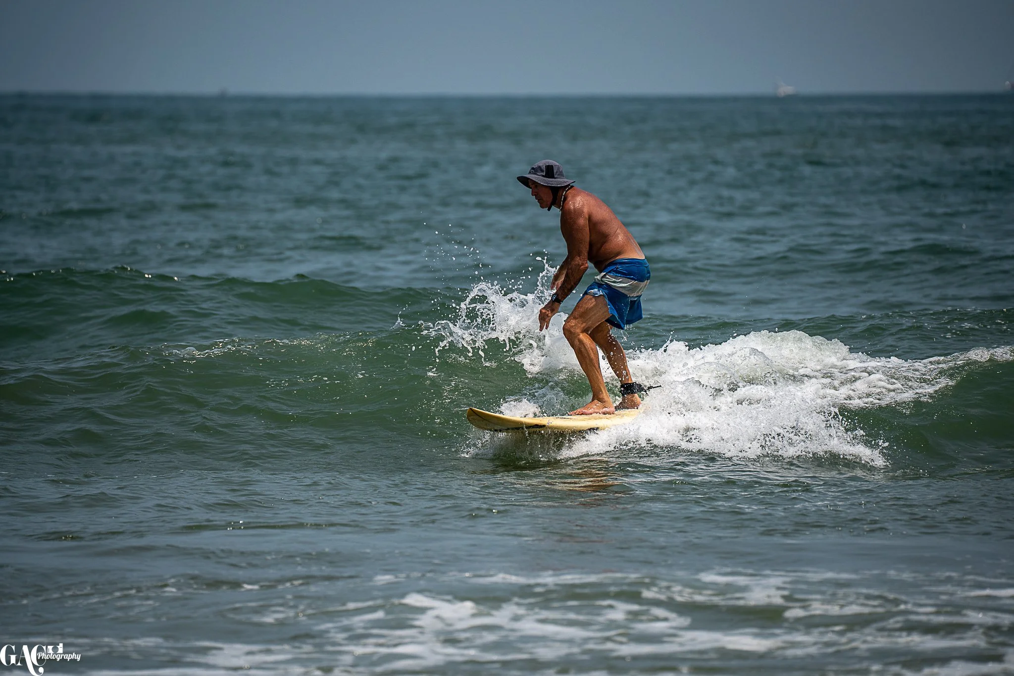 A man surfing on a wave in the ocean, wearing blue shorts and a hat.