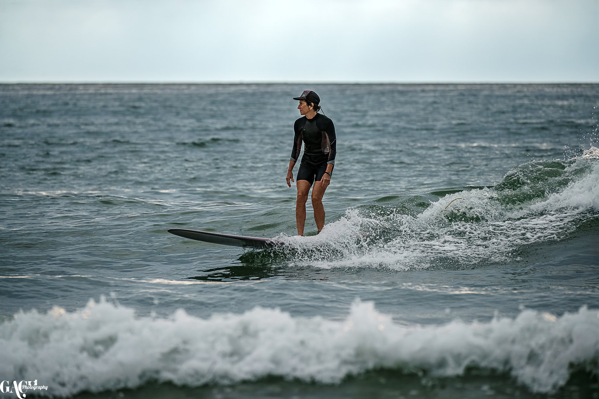 A woman in a black wetsuit and cap surfboarding on an ocean wave during cloudy weather.