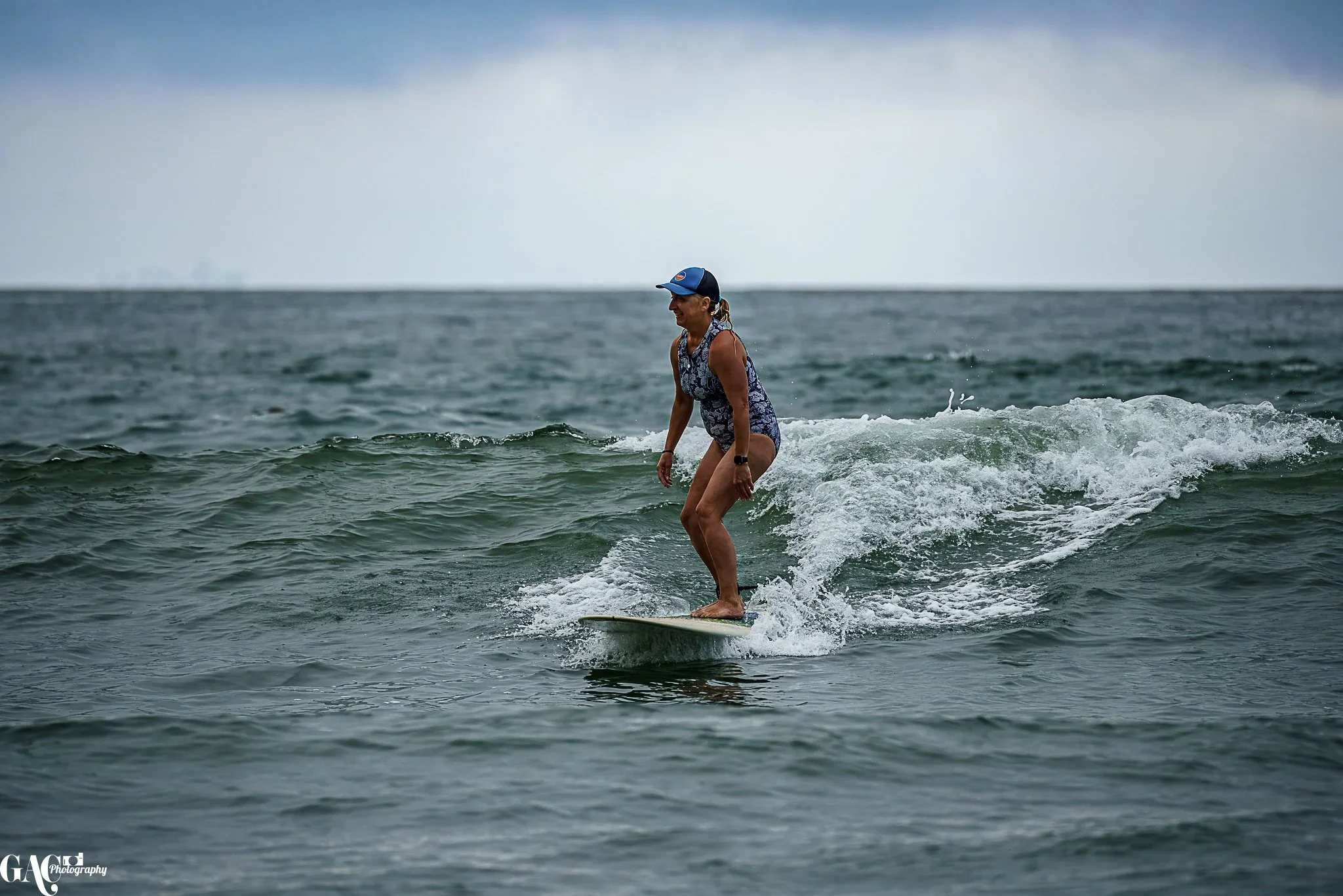 A woman surfing on the ocean waves under a cloudy sky, wearing a blue swimsuit, cap, and watch.