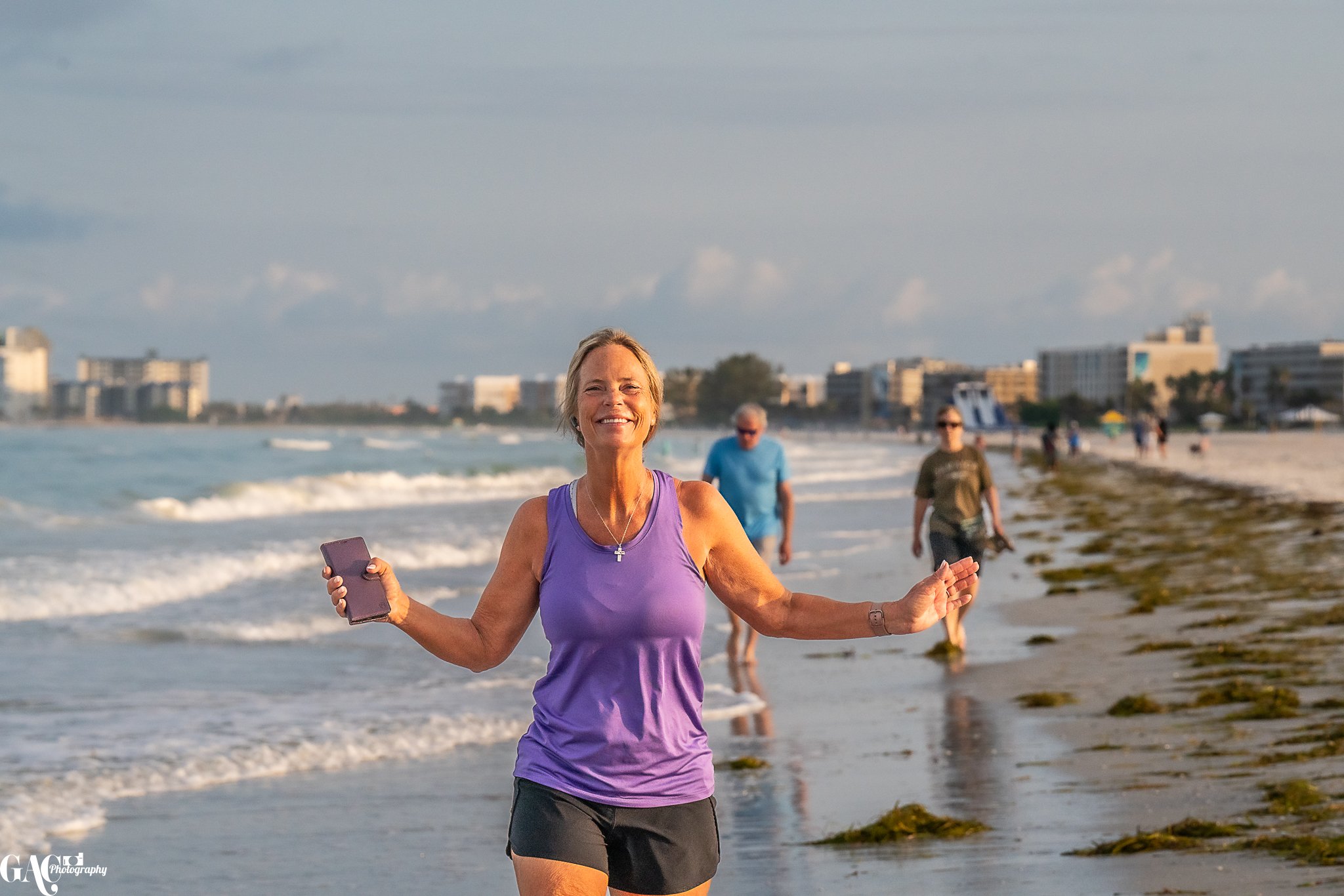 A woman in a purple tank top and black shorts walking on a beach, smiling and holding a phone. The beach has scattered seaweed and there are people walking in the background. Buildings are visible in the distance.