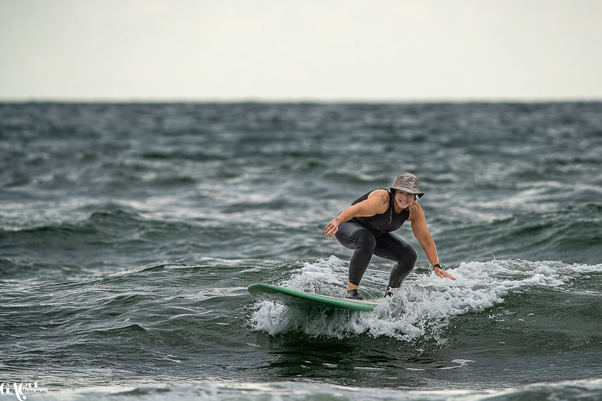 A woman surfing on a small wave in the ocean, wearing a black sleeveless top, black leggings, and a gray bucket hat, smiling and balancing on her surfboard.