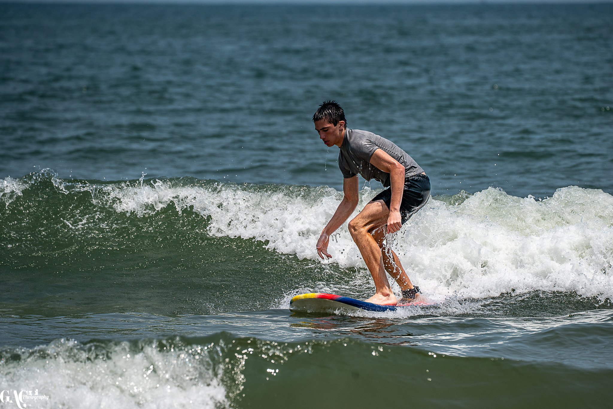 A young man surfing on a small wave at the beach, wearing a gray t-shirt and black shorts.
