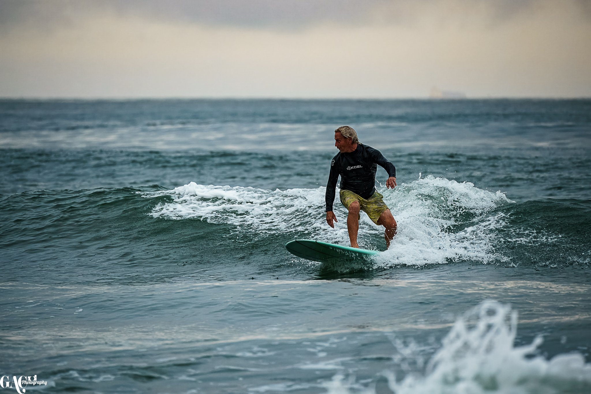 A man surfing on a small wave in the ocean, wearing a dark long-sleeve shirt and camouflage shorts.