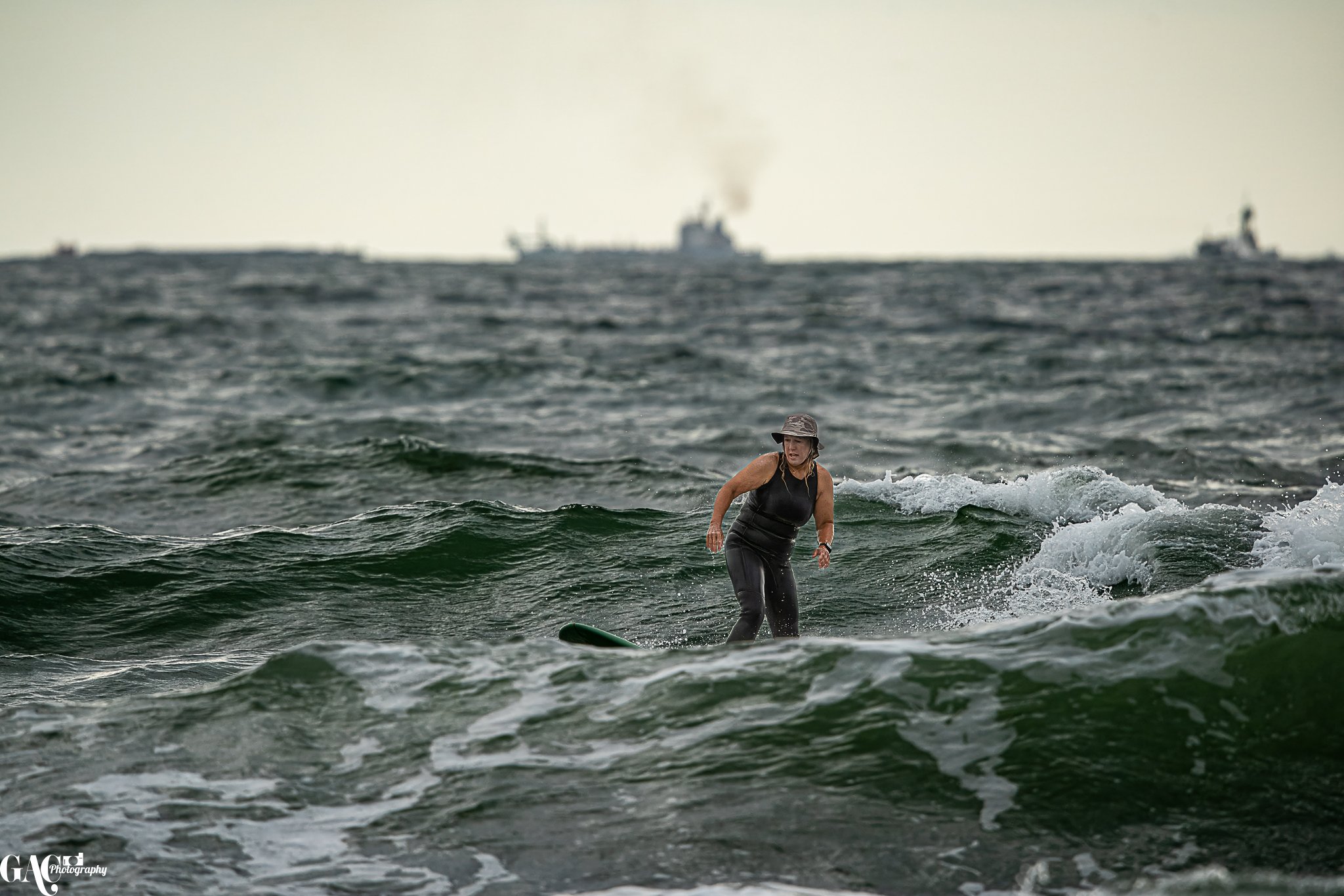 Woman in black wetsuit and gray bucket hat surfing on a wave in the ocean, with ships visible in the background.