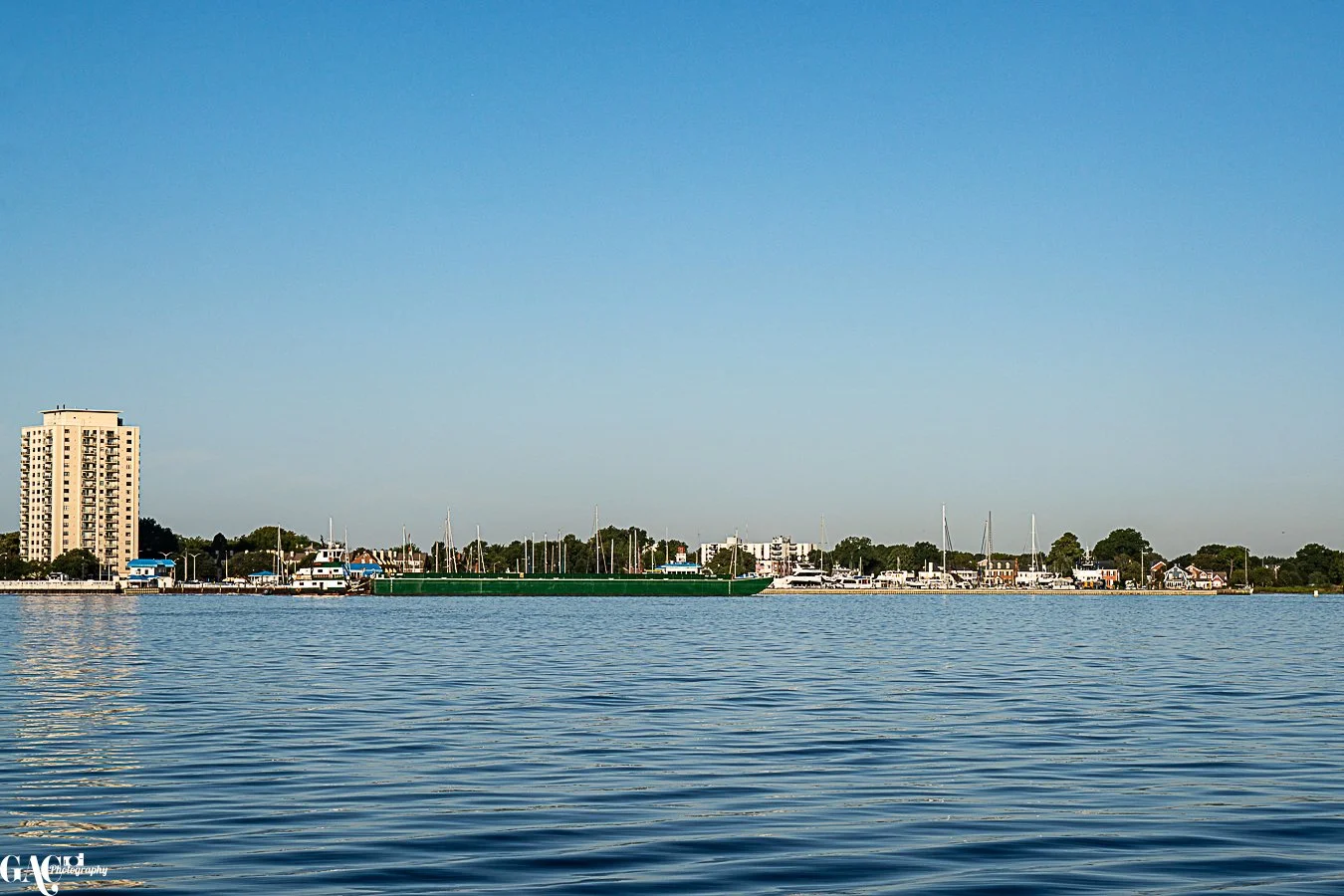 A body of water with a cityscape in the background, including boats and a tall building under a clear blue sky.