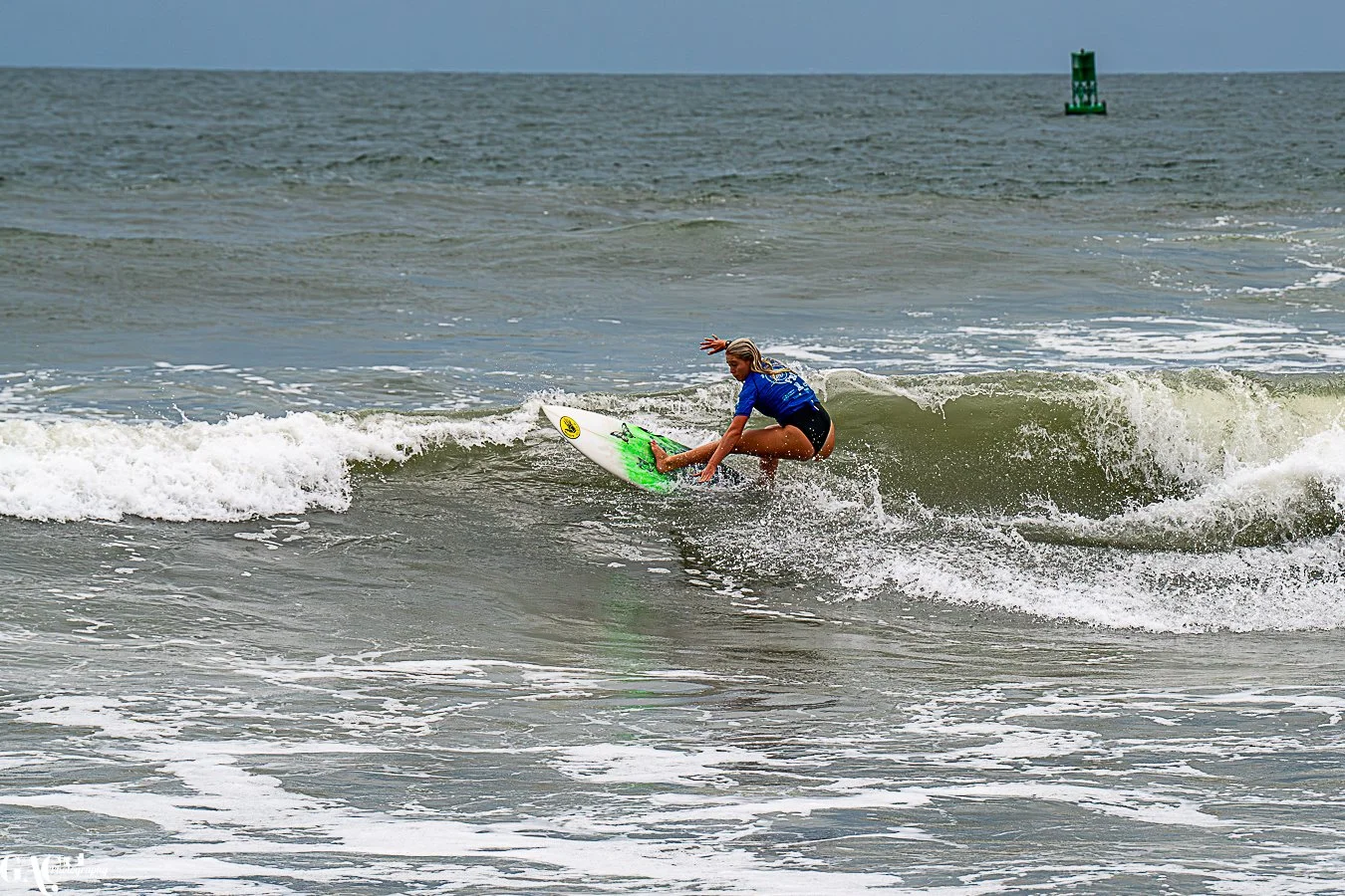 A female surfer riding a small wave near the shore with a green buoy in the distance.