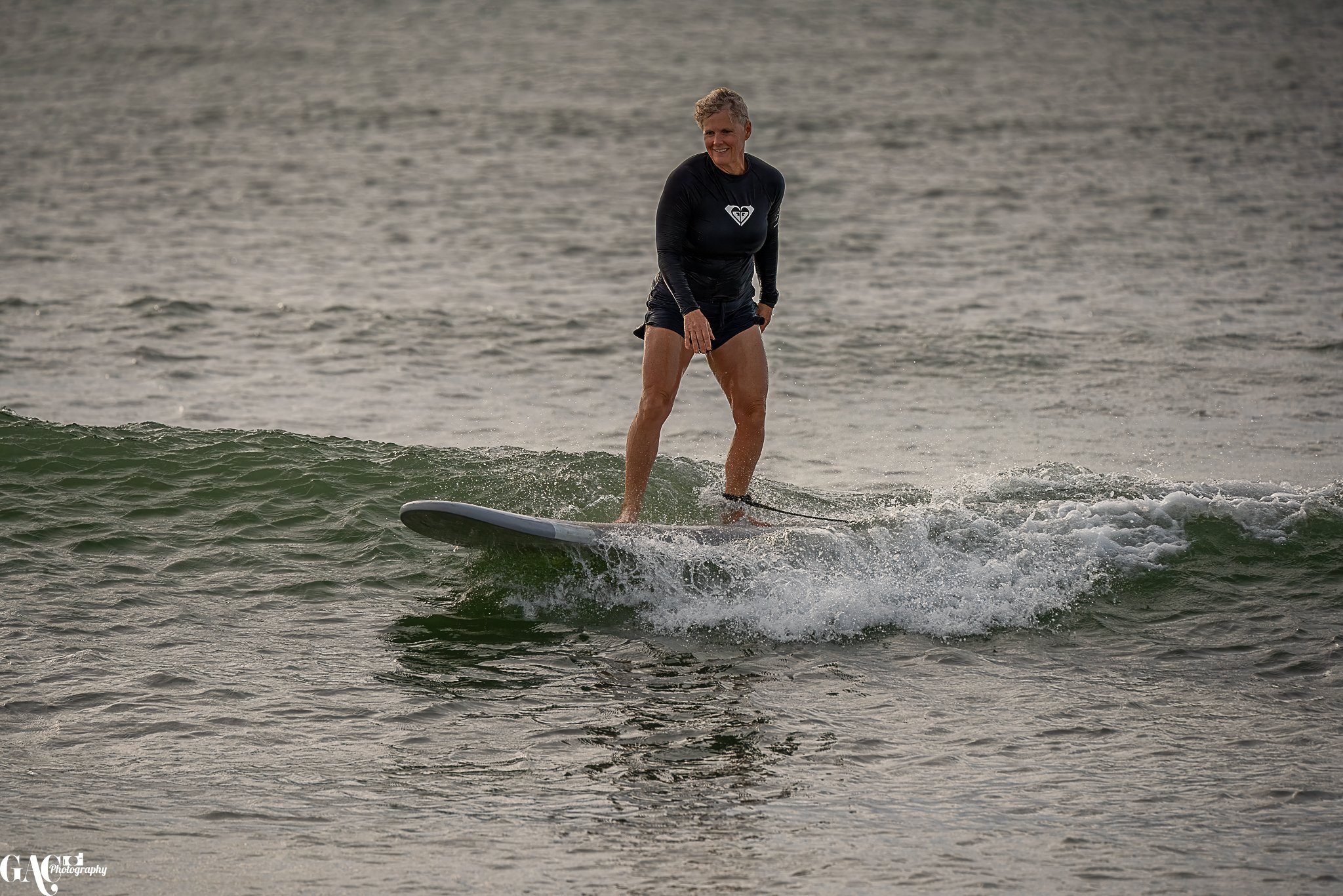 A woman with short hair wearing a black long-sleeve rash guard and black shorts surfing on a small wave.