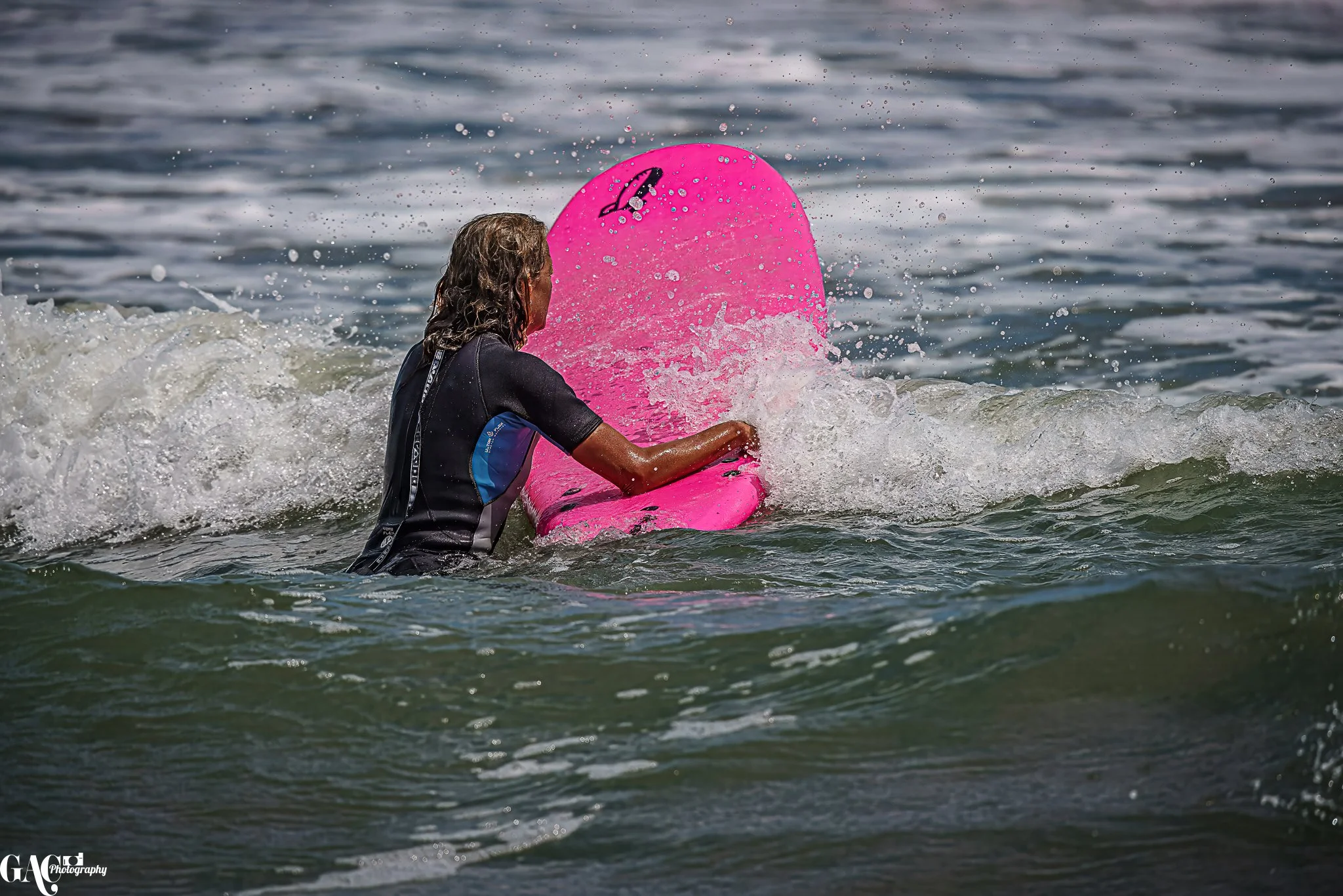 Person with long hair wearing a wetsuit, paddling a pink surfboard in the ocean waves.