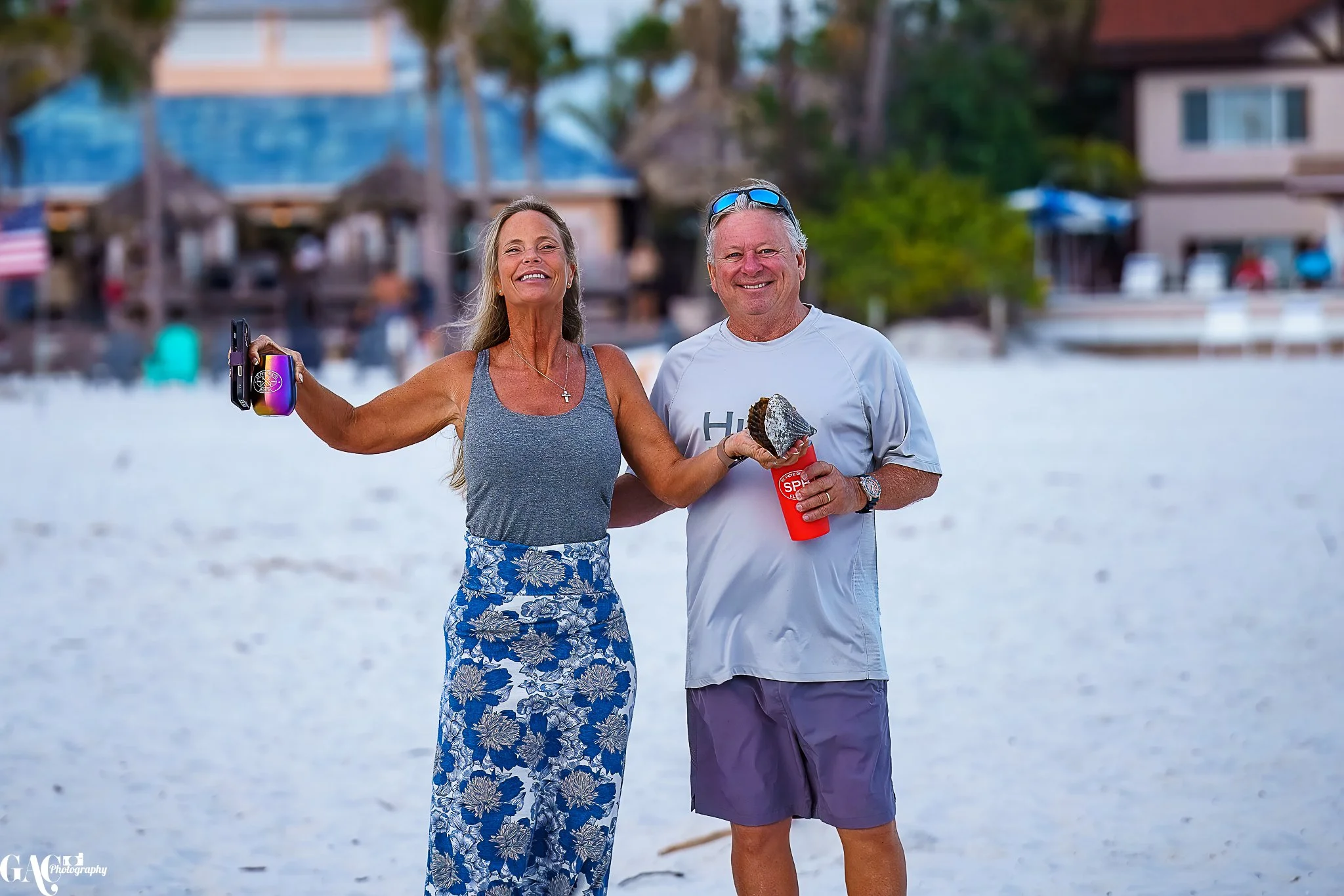 A joyful couple posing on a beach, the woman holding a phone with a colorful case, and the man holding a red cup and a large shell. Palm trees and buildings are blurred in the background.