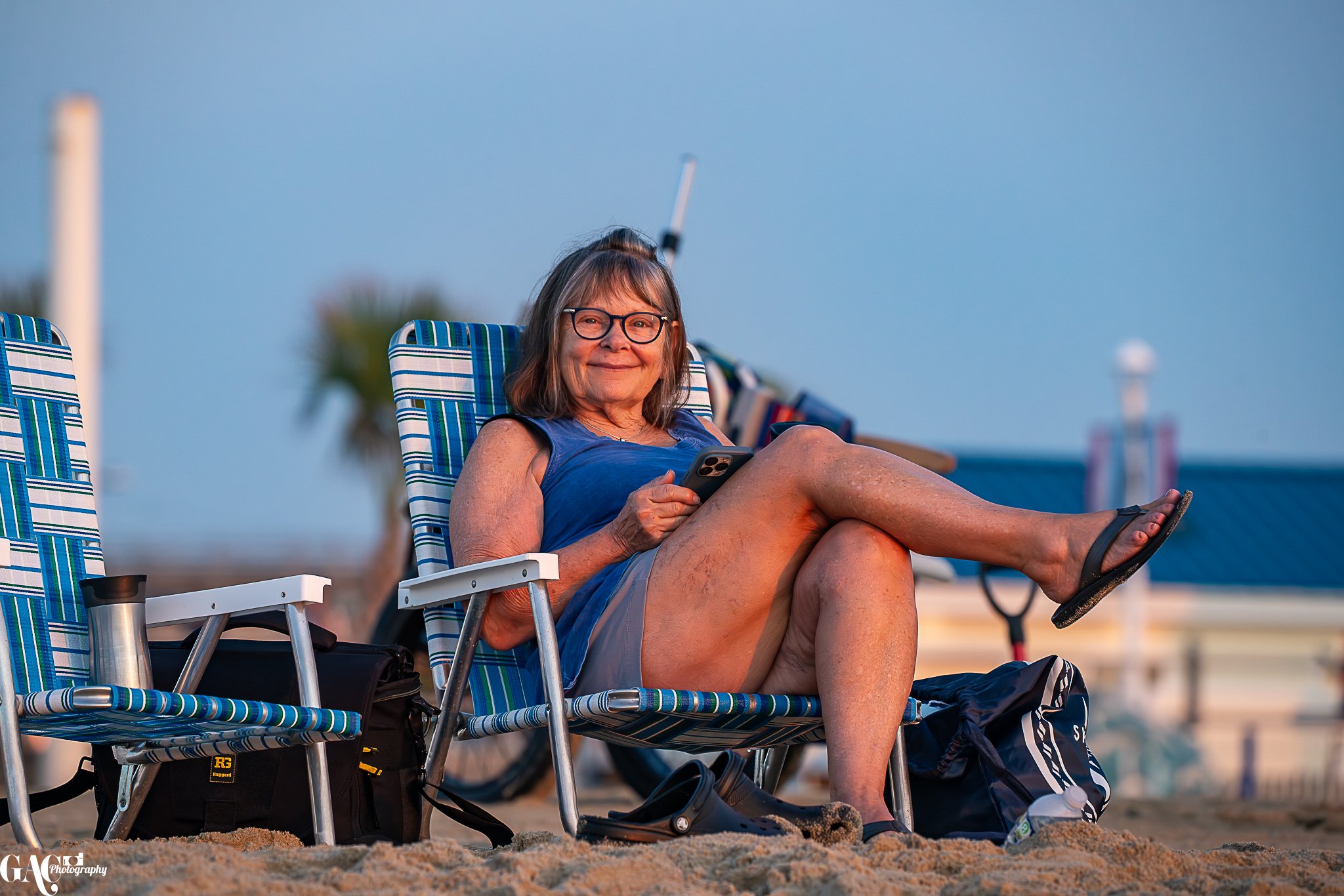 Older woman sitting on a beach chair, smiling and holding a smartphone, with beach bags and a drink nearby, in the evening or late afternoon.